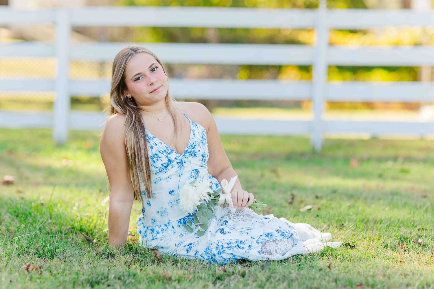 Young blonde woman in blue floral dress sitting on green grass near a white wooden fence.