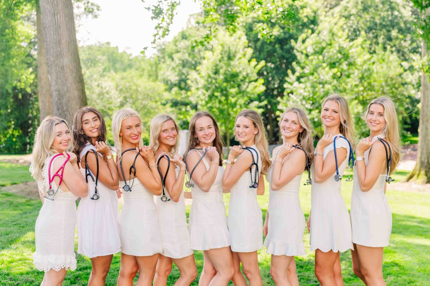 Group of women in white dresses holding stethoscopes outdoors, celebrating nursing school milestone.