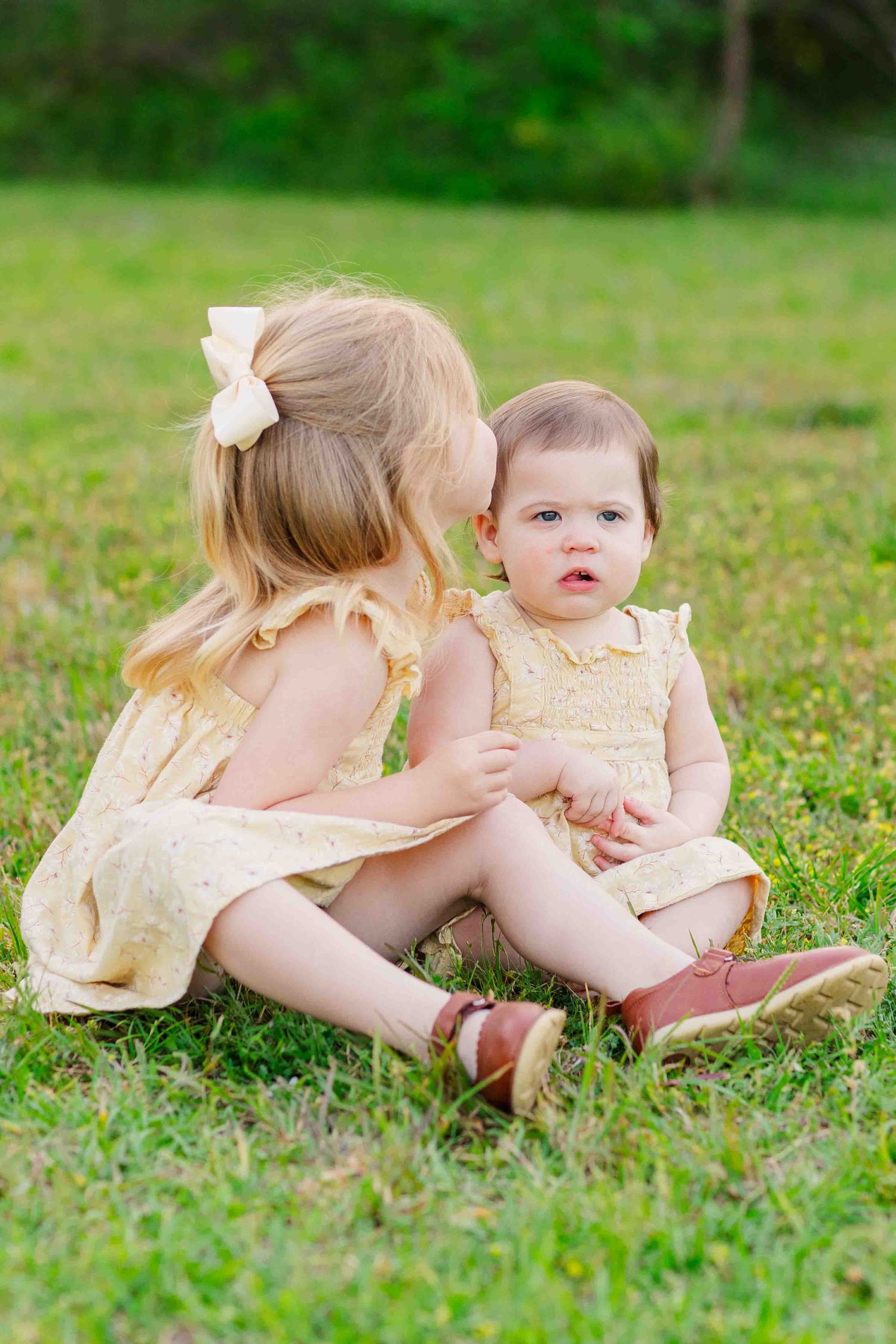 Two young sisters in matching yellow dresses sitting on grass as older sibling kisses the baby.
