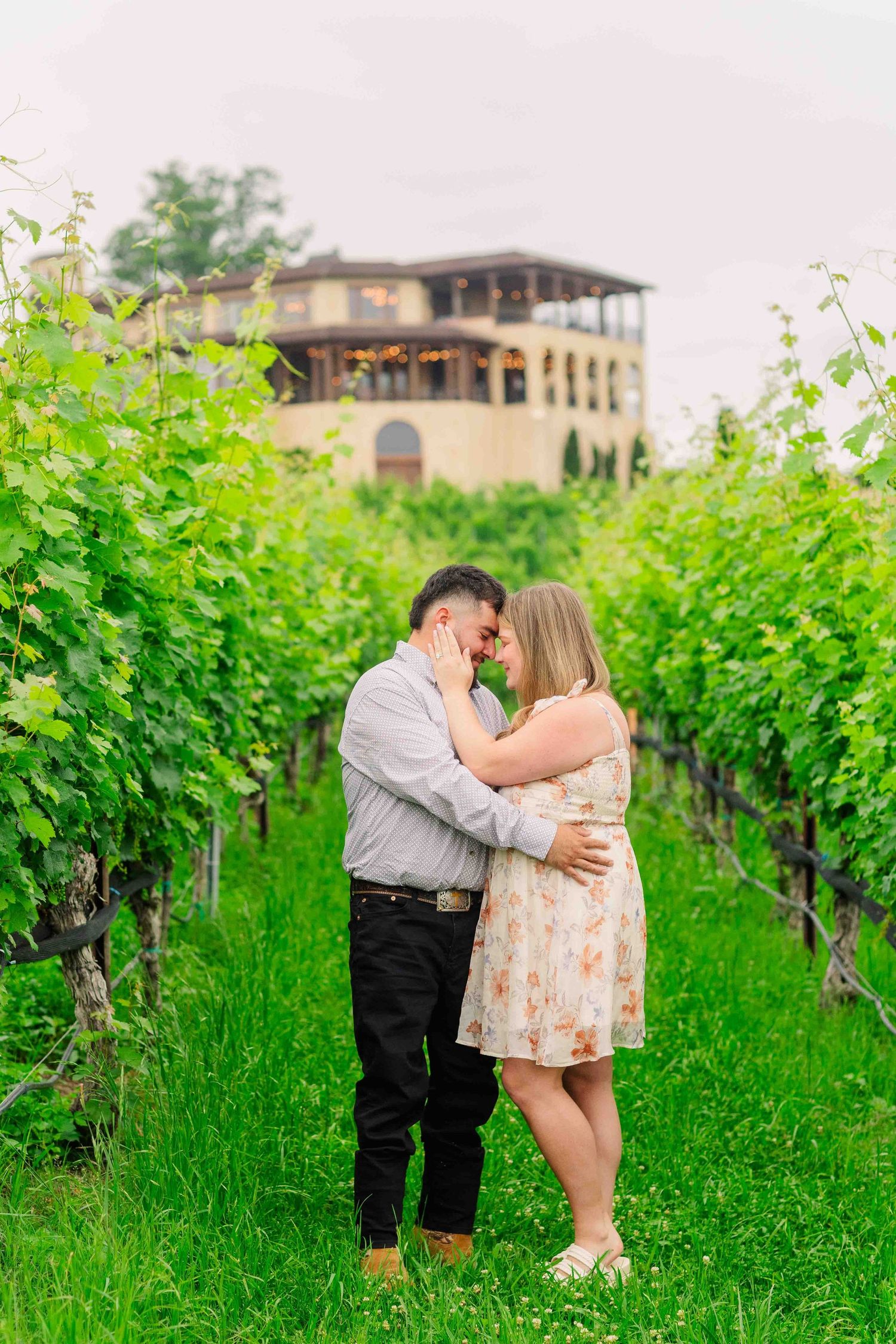 Couple embracing romantically between lush green vineyard rows with elegant winery building in background.