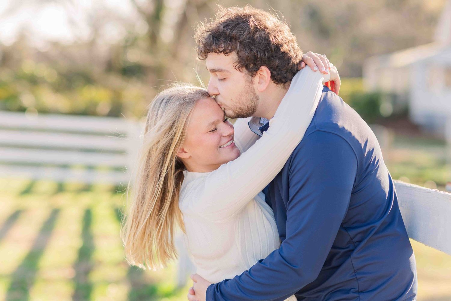Man in navy jacket kissing woman on cheek while embracing her outdoors in warm sunlight.