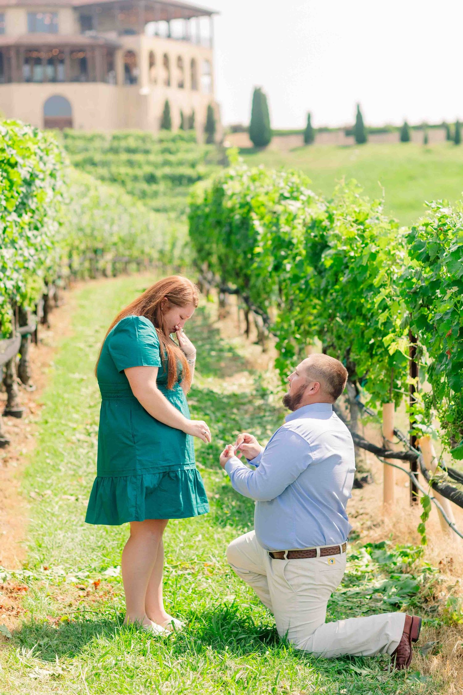 Man proposing on one knee to woman in green dress in a lush vineyard with winery building behind.