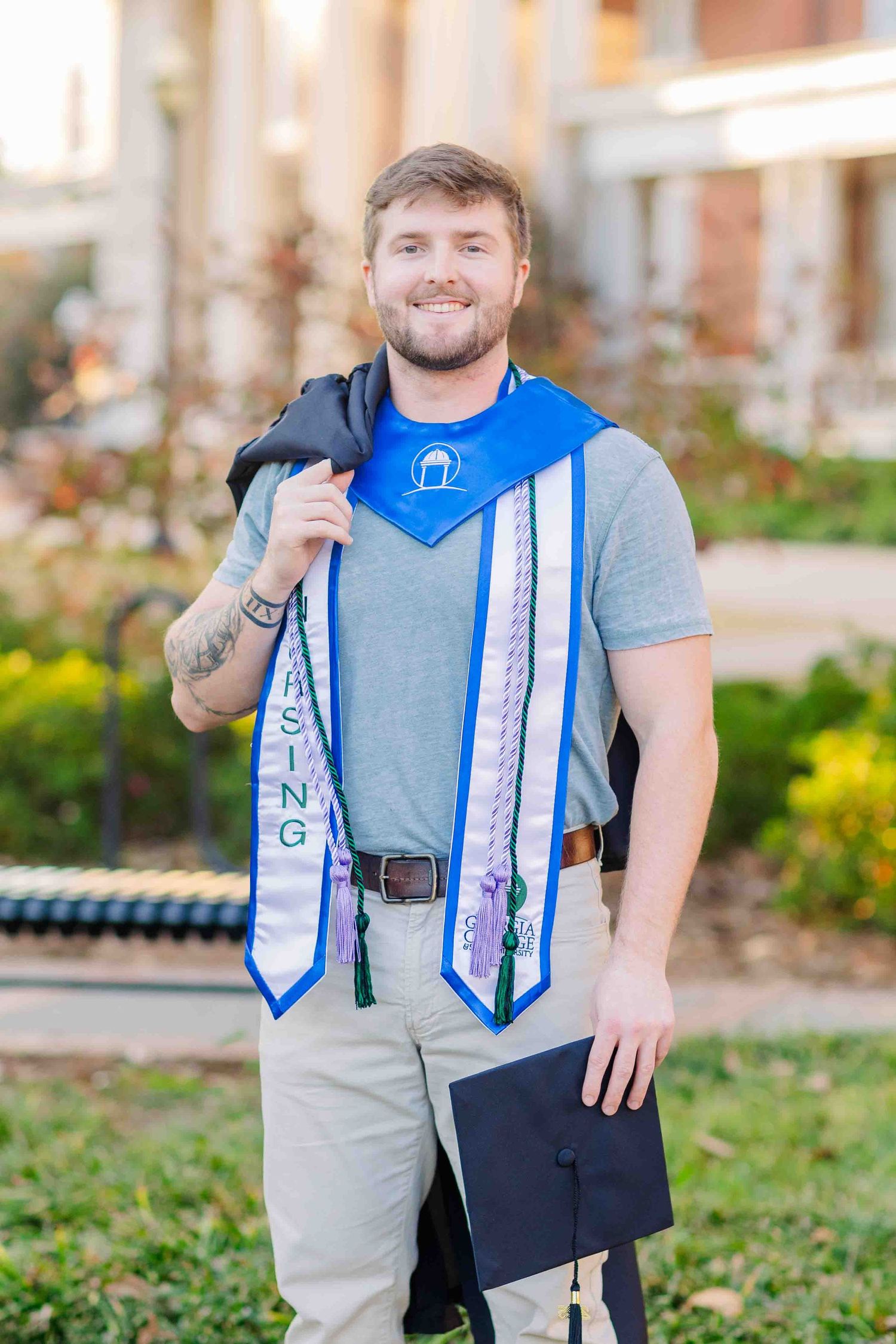 Male nursing graduate smiles holding cap and gown with blue stole and multiple honor cords.