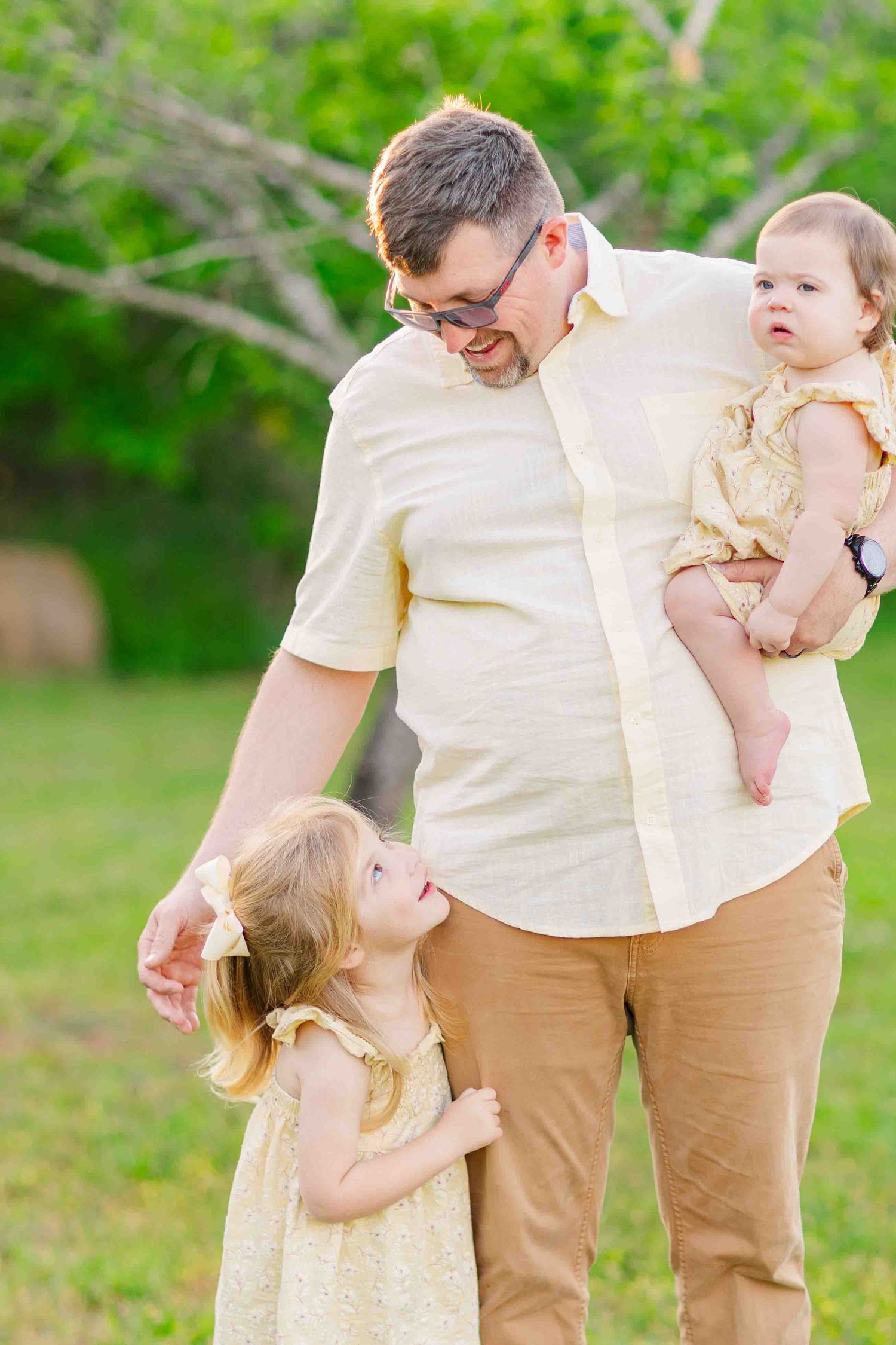 Cheerful father with glasses holds baby girl while toddler daughter hugs his leg outdoors.