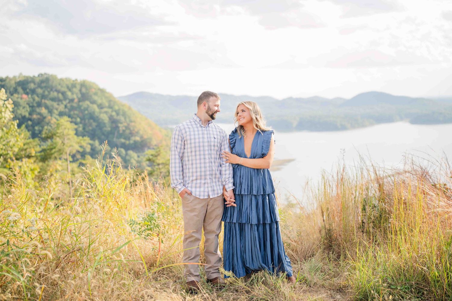 Couple holding hands and smiling at each other in golden field with lake and mountains behind them.