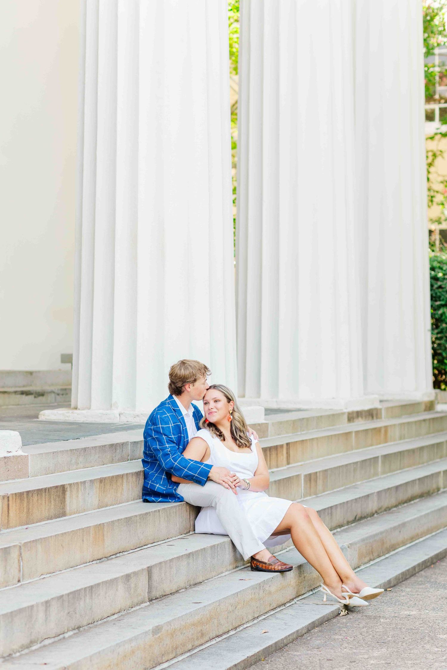Couple sitting on stone steps near white columns, man kissing womans cheek during engagement session.