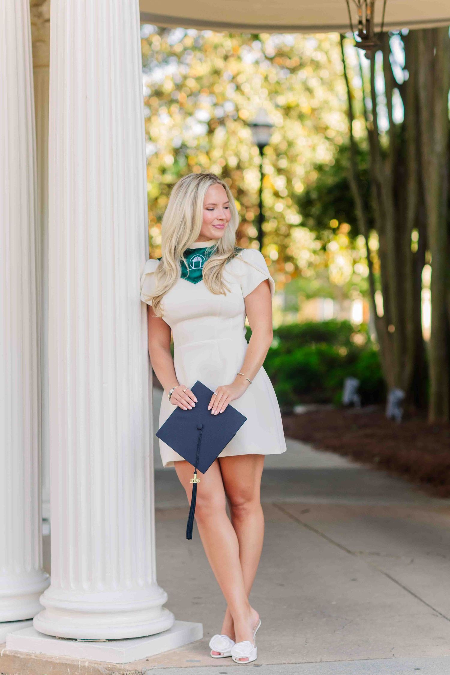 Blonde woman in white dress holding navy graduation cap poses by white column outdoors.