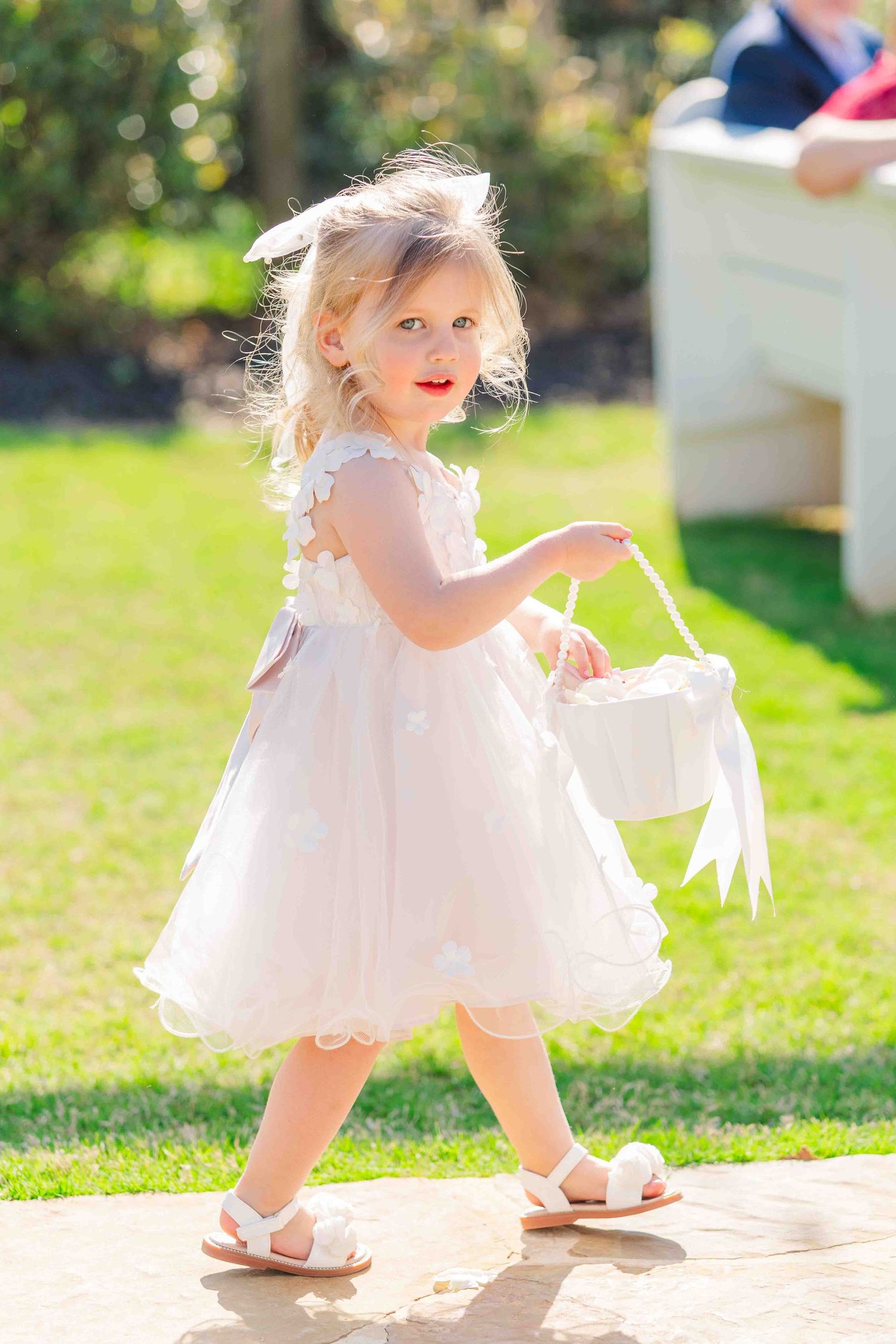 Adorable blonde flower girl in white dress carries basket down outdoor wedding aisle.