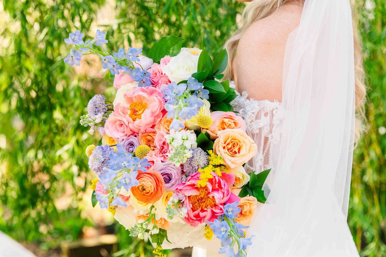Bride holding vibrant colorful wedding bouquet with pink, orange, and blue flowers outdoors.