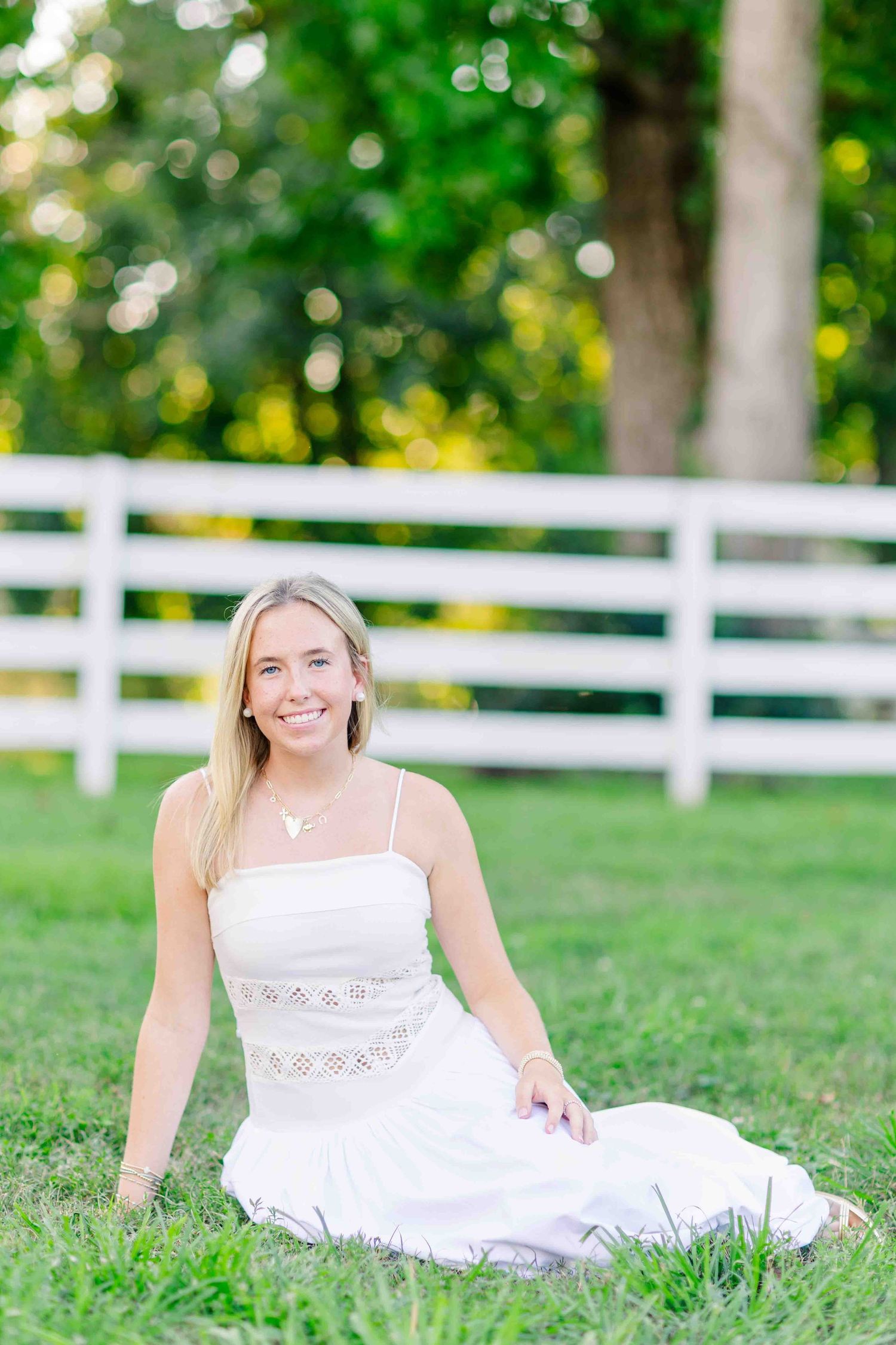 Smiling blonde woman in white sundress sitting on green grass near white fence outdoors.