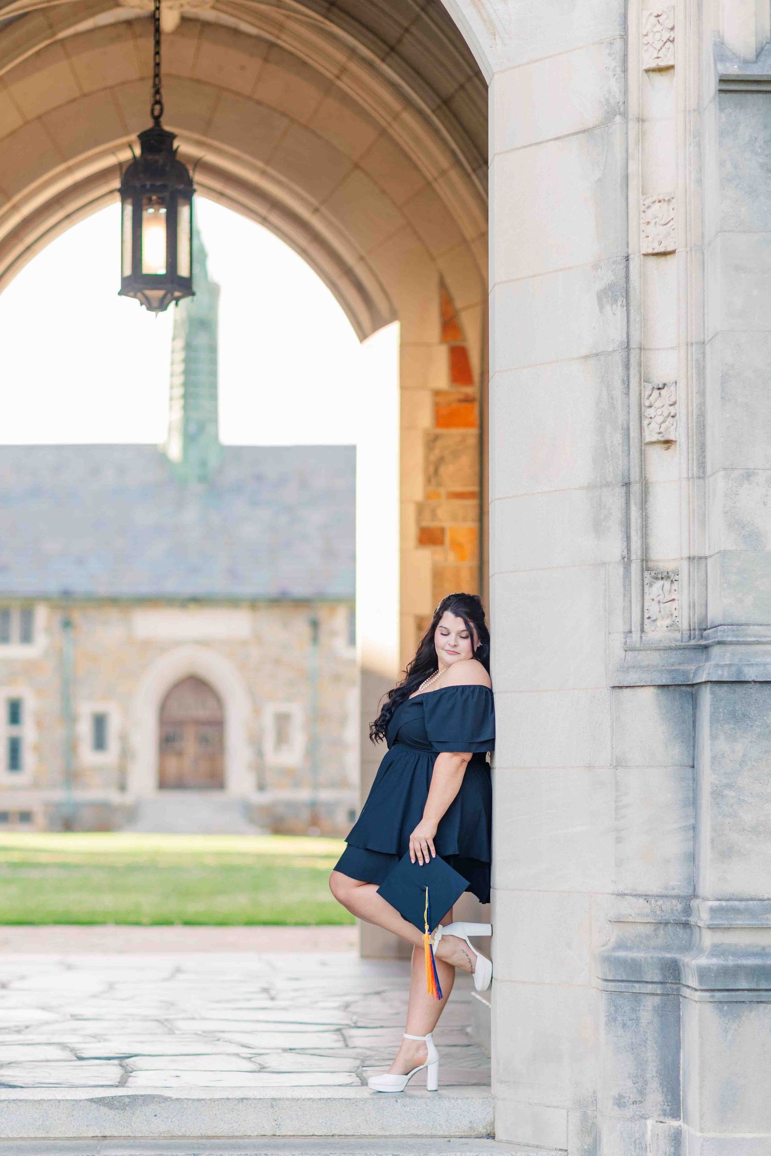 Graduate in black off-shoulder dress holding cap under stone archway at university campus.