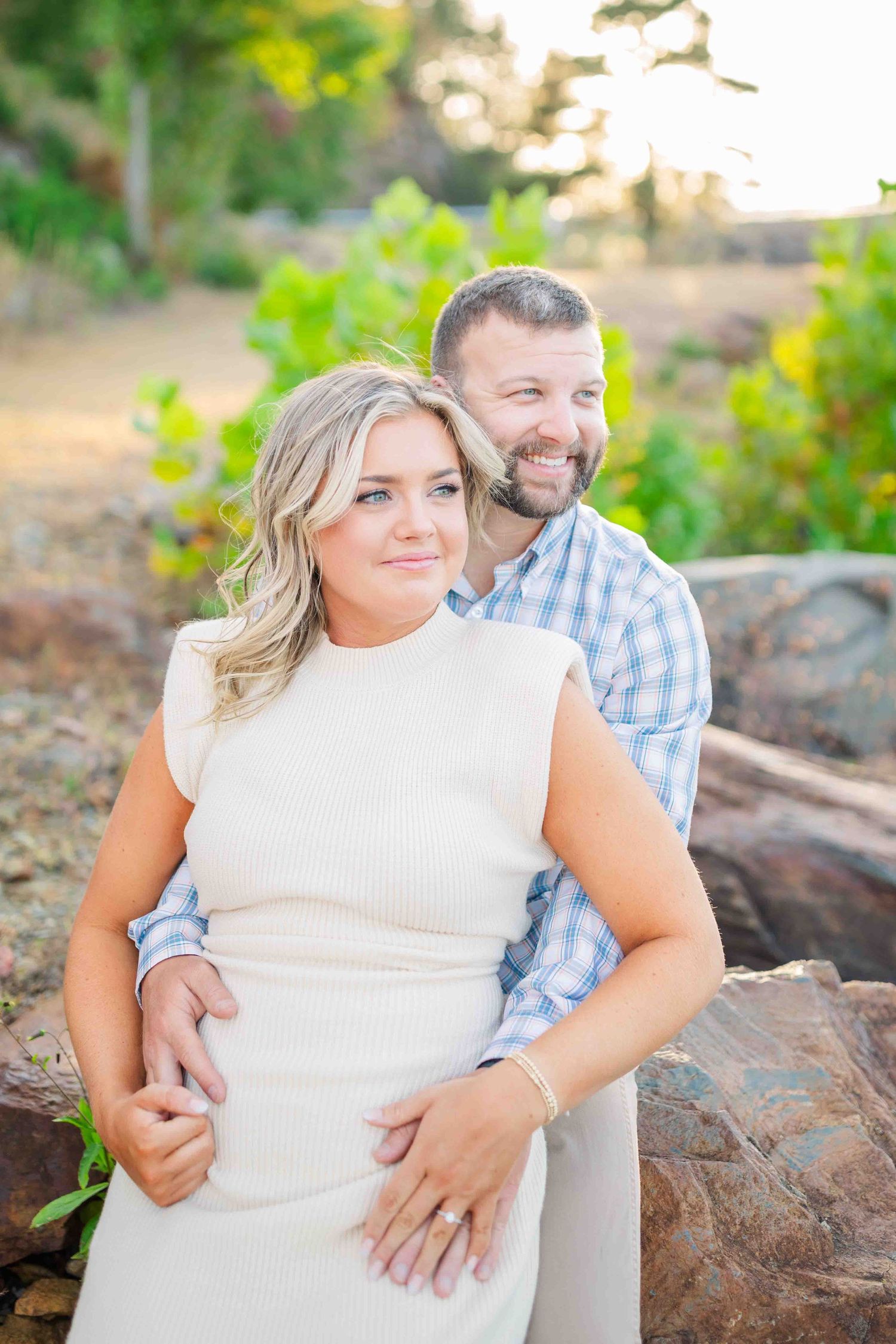 Couple posing outdoors at golden hour, woman in cream dress with man in plaid shirt embracing her from behind near rocks.