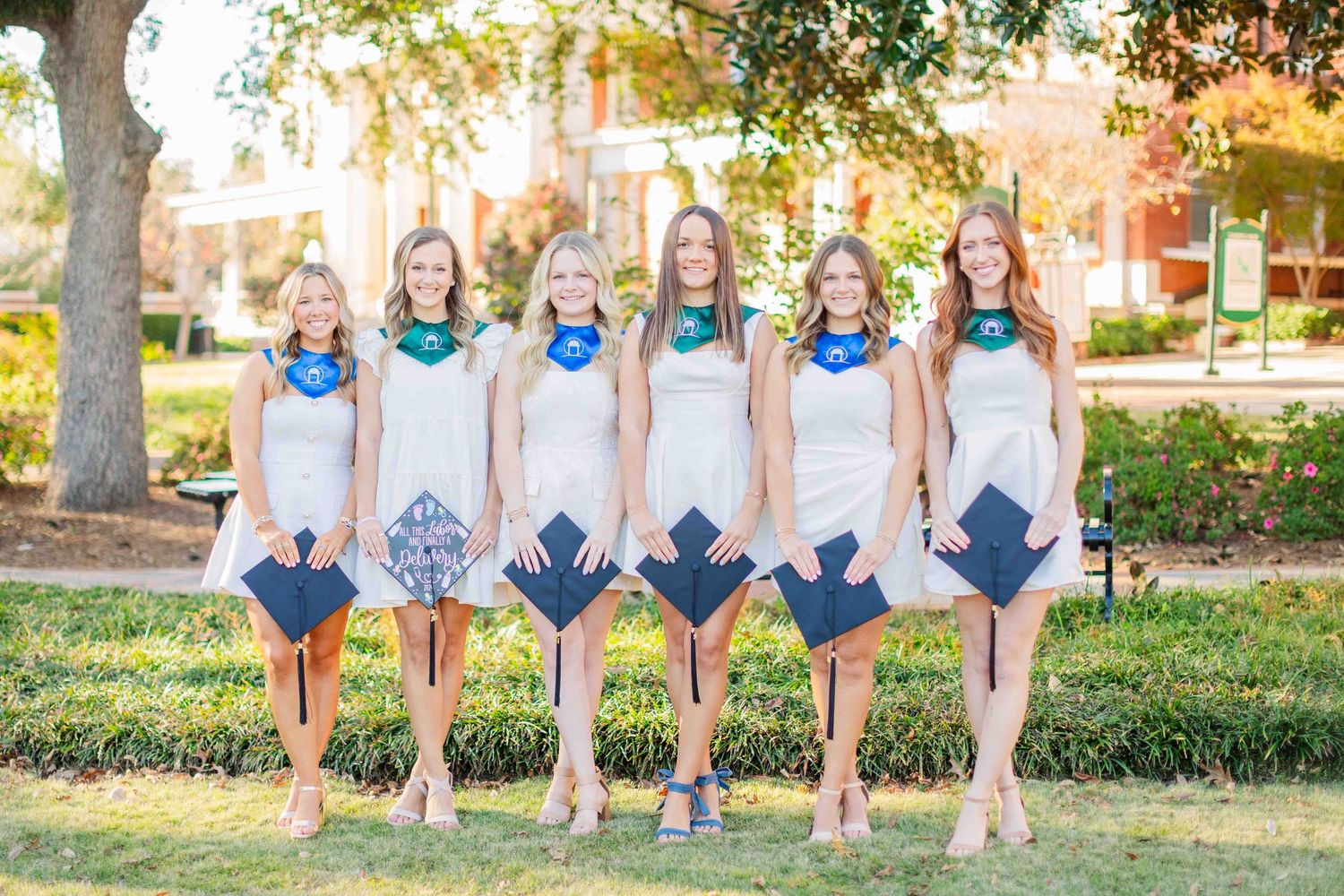 Six young women in white dresses holding blue graduation caps outdoors on a sunny college campus.