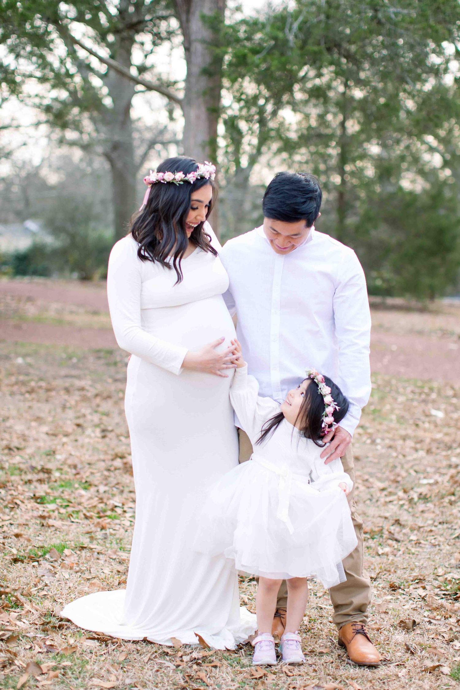 Expecting family in white outfits with flower crowns posing outdoors in wooded setting.