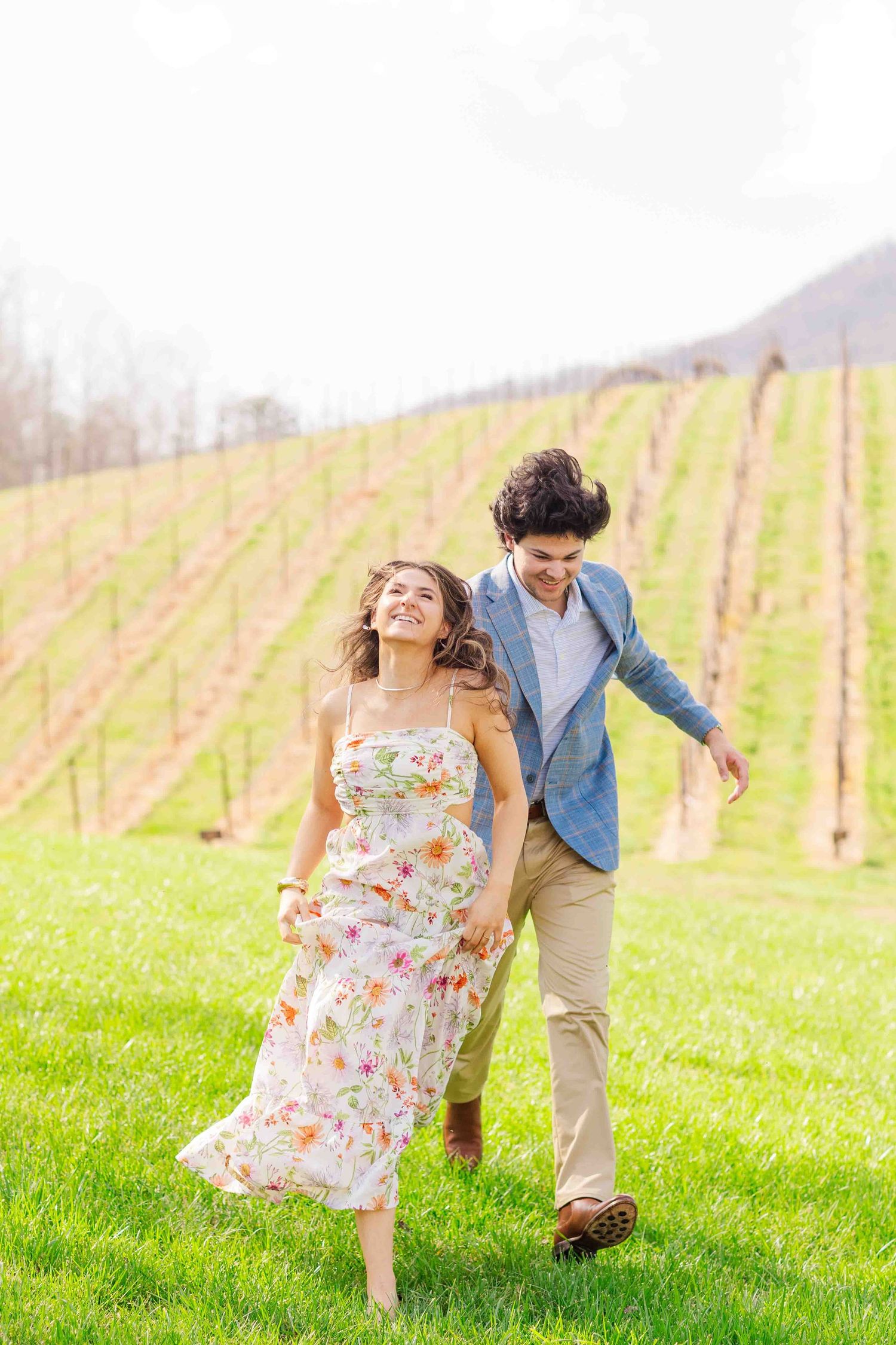 Joyful couple holding hands and running through green grass with vineyard rows and mountain behind them.