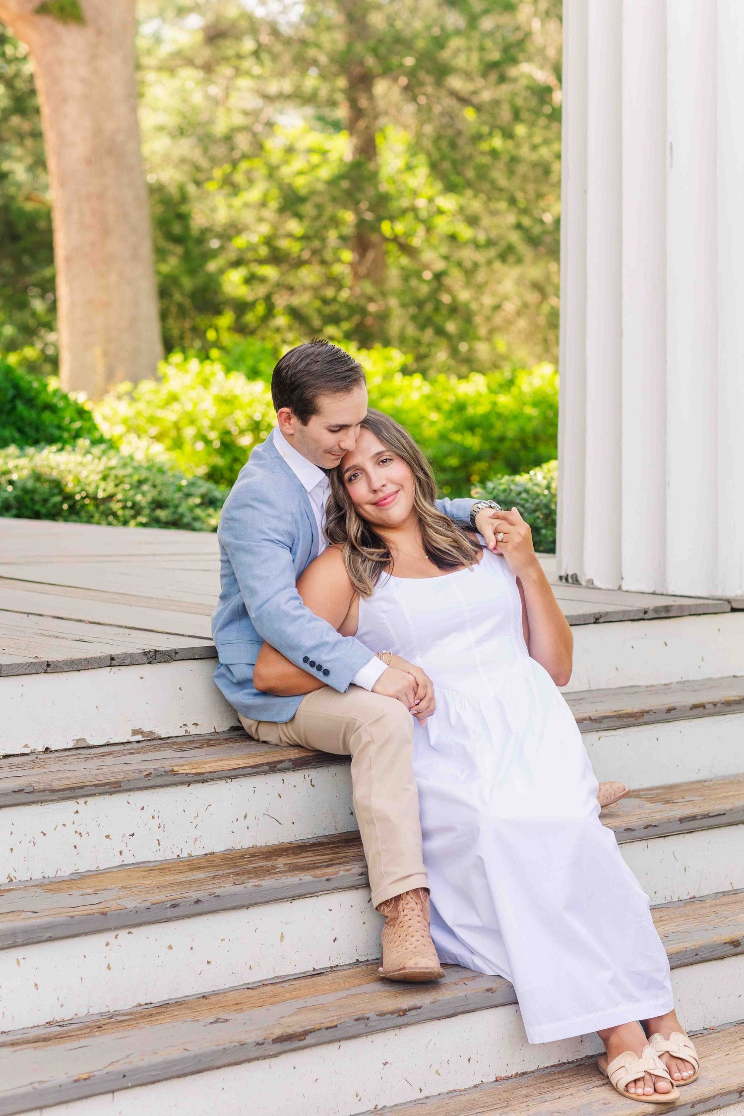 Couple sitting on white colonial steps, woman in white dress leaning against man in blue blazer and khaki pants.