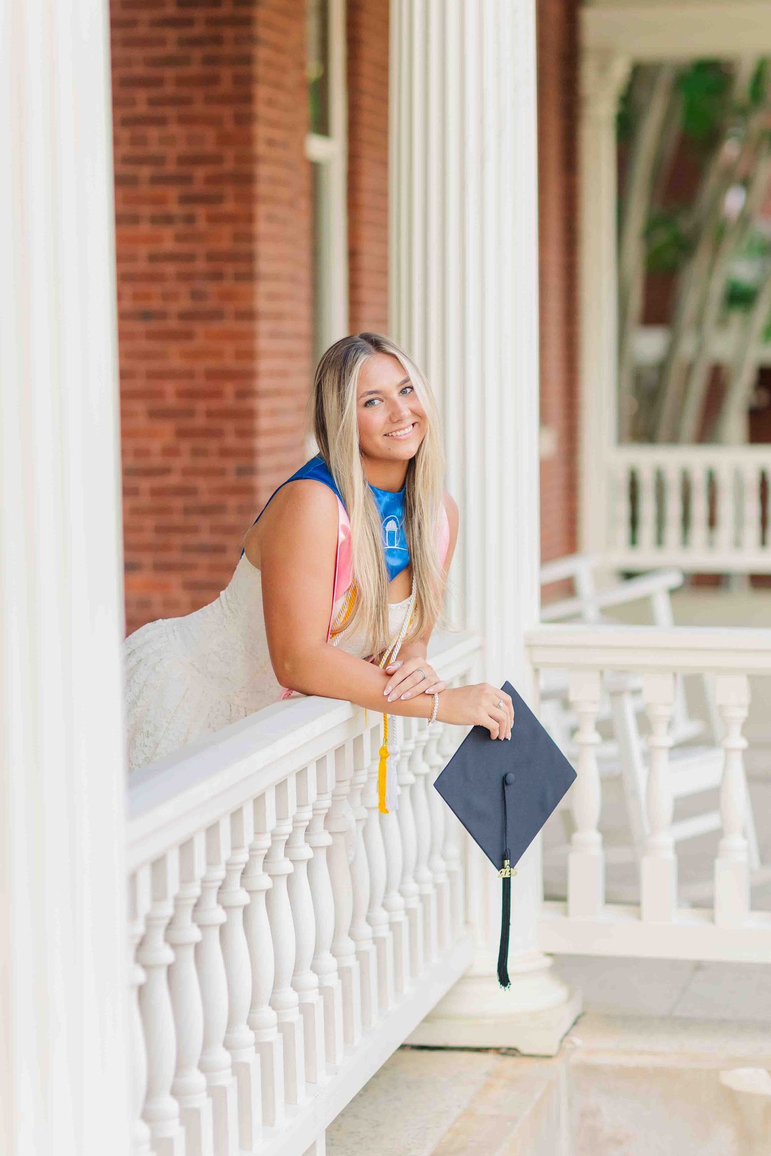 Smiling blonde graduate leans on white porch railing holding black cap with green tassel.