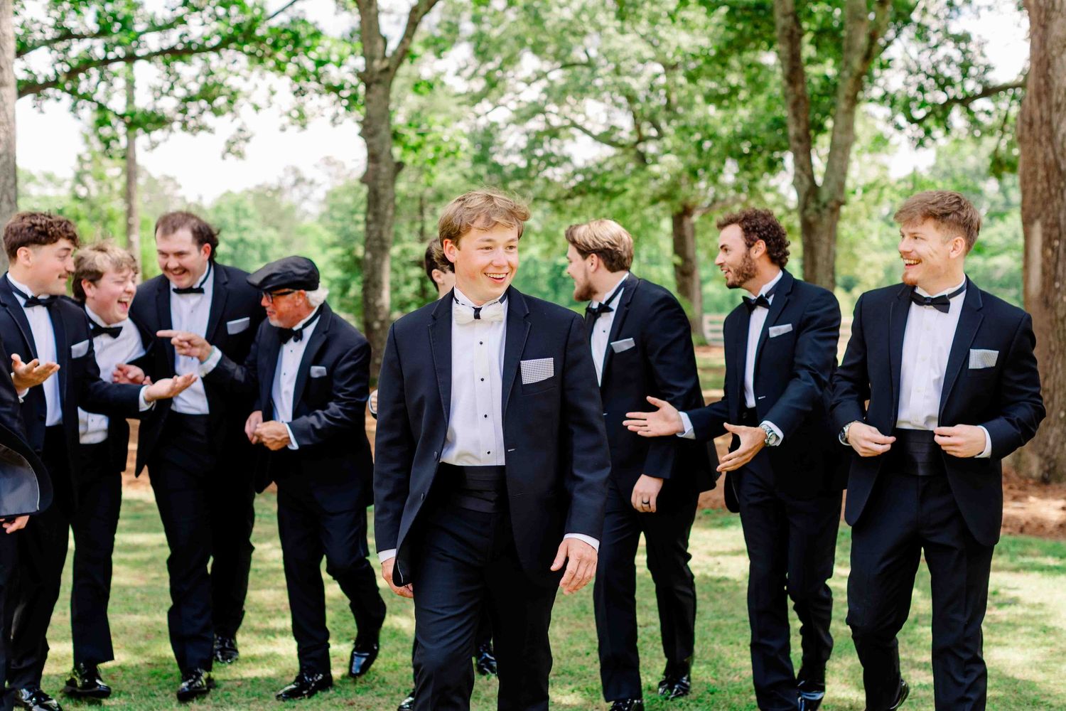 Groom and groomsmen in black tuxedos laughing outdoors under green trees.