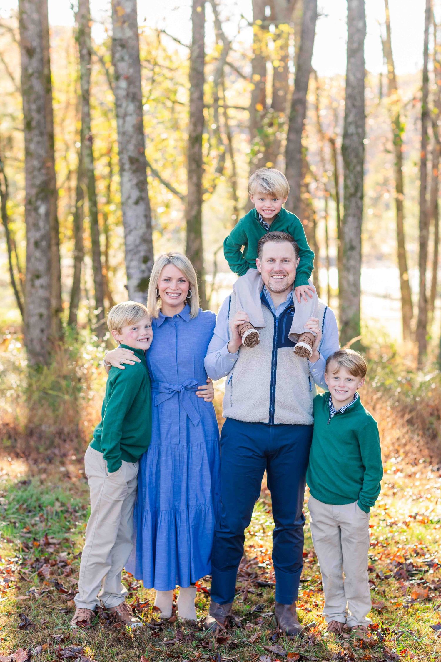 Smiling family of five in green and blue coordinating outfits posing together in fall forest setting.