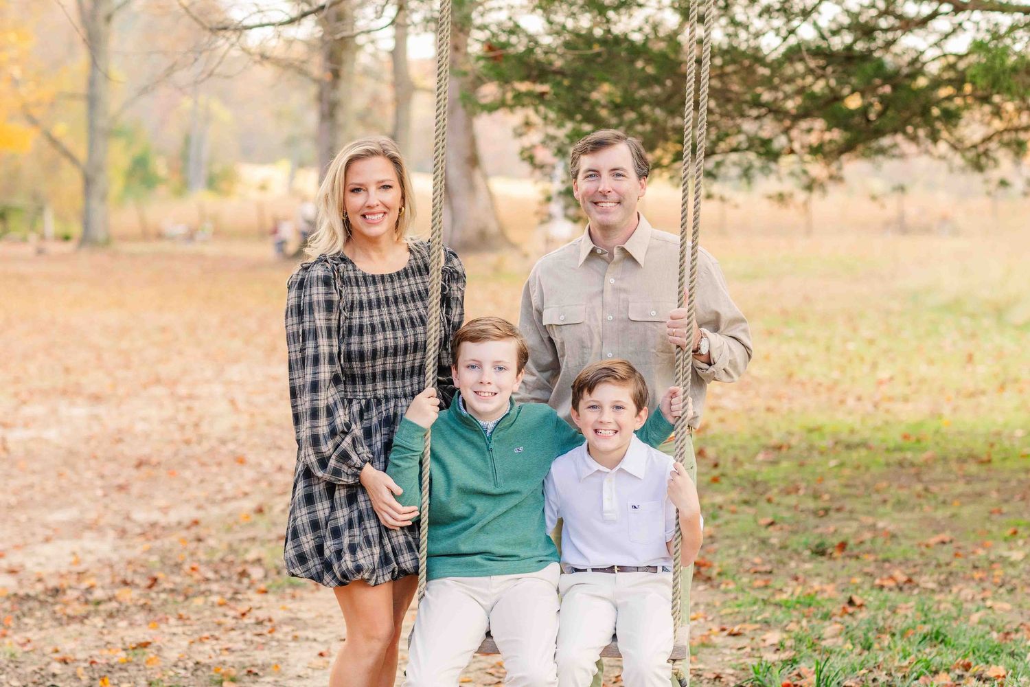 Smiling family of four posing near rustic swing in golden autumn park, boys in green polo shirts.
