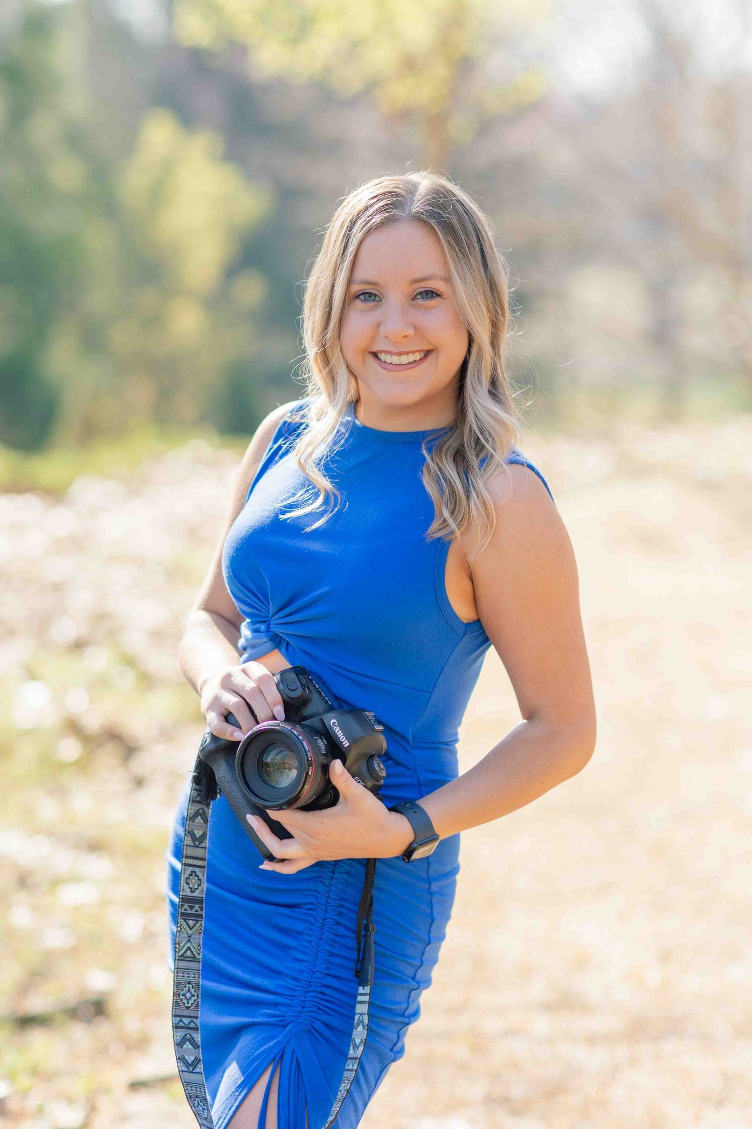 Smiling blonde photographer in blue dress holding camera in sunny outdoor setting.