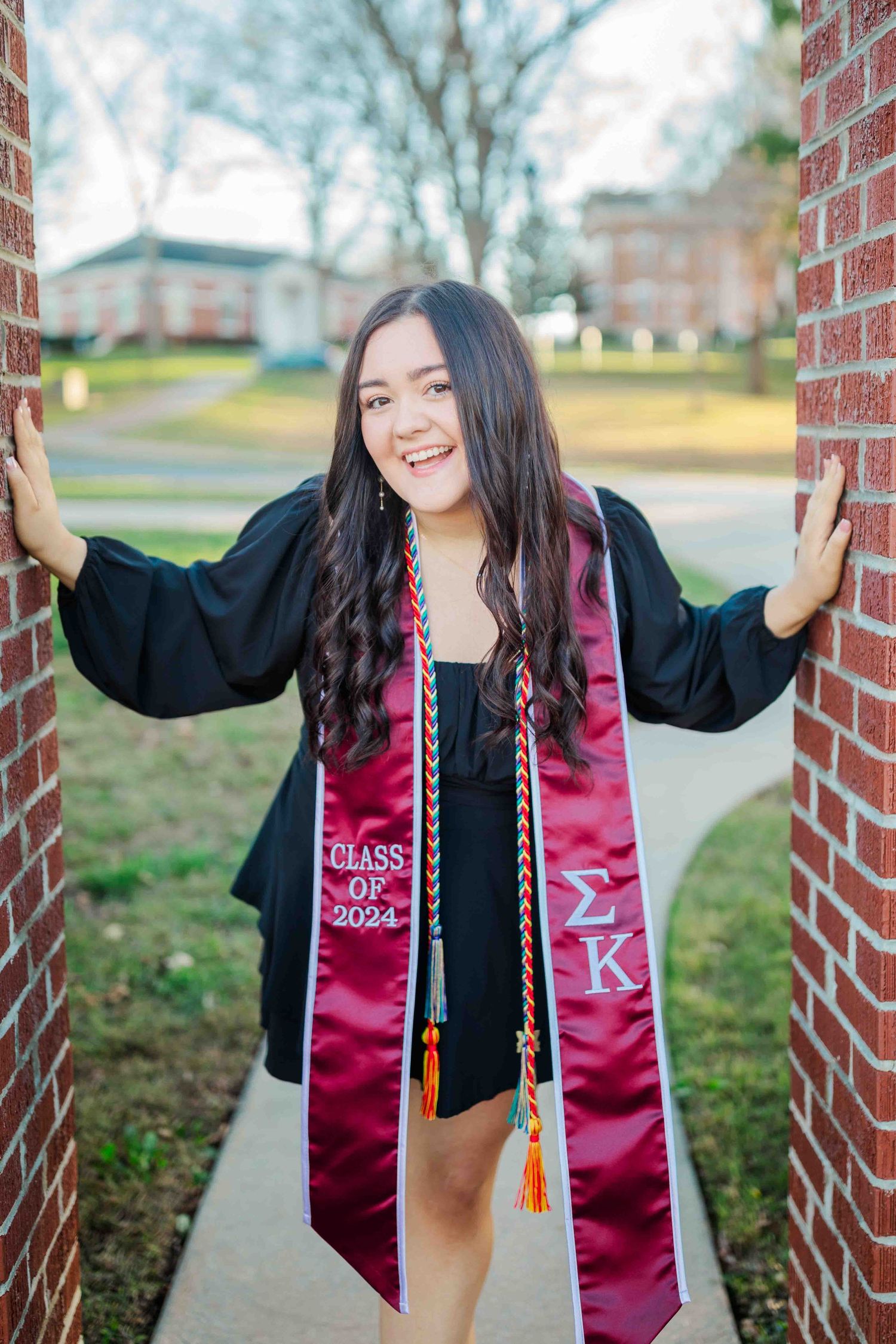 Smiling college graduate in black gown wearing Sigma Kappa Class of 2024 stole poses between brick columns on campus.