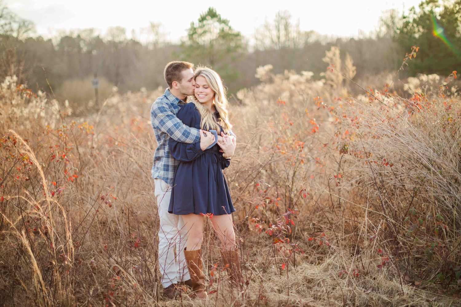 Couple embracing in golden autumn field, man in plaid shirt kissing woman in navy blue dress.