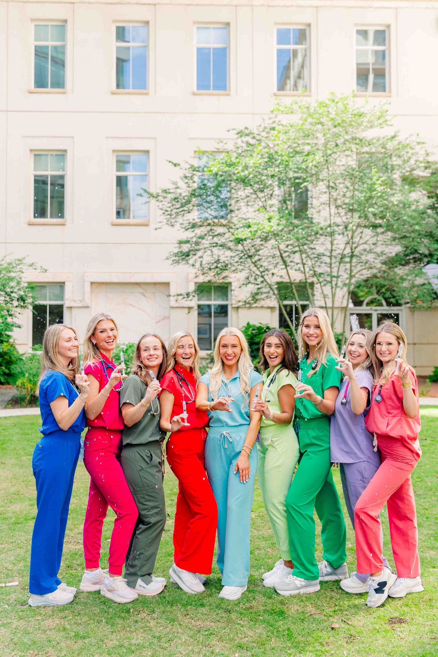 Group of nursing students in colorful scrubs posing with stethoscopes outside university building.