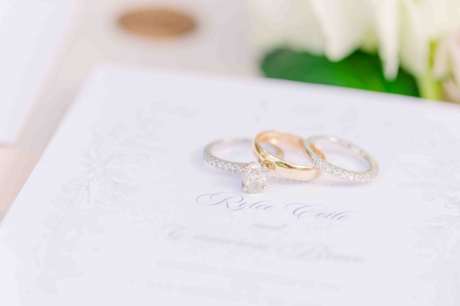 Three elegant wedding rings featuring a diamond solitaire and two bands displayed on a white wedding invitation with florals.