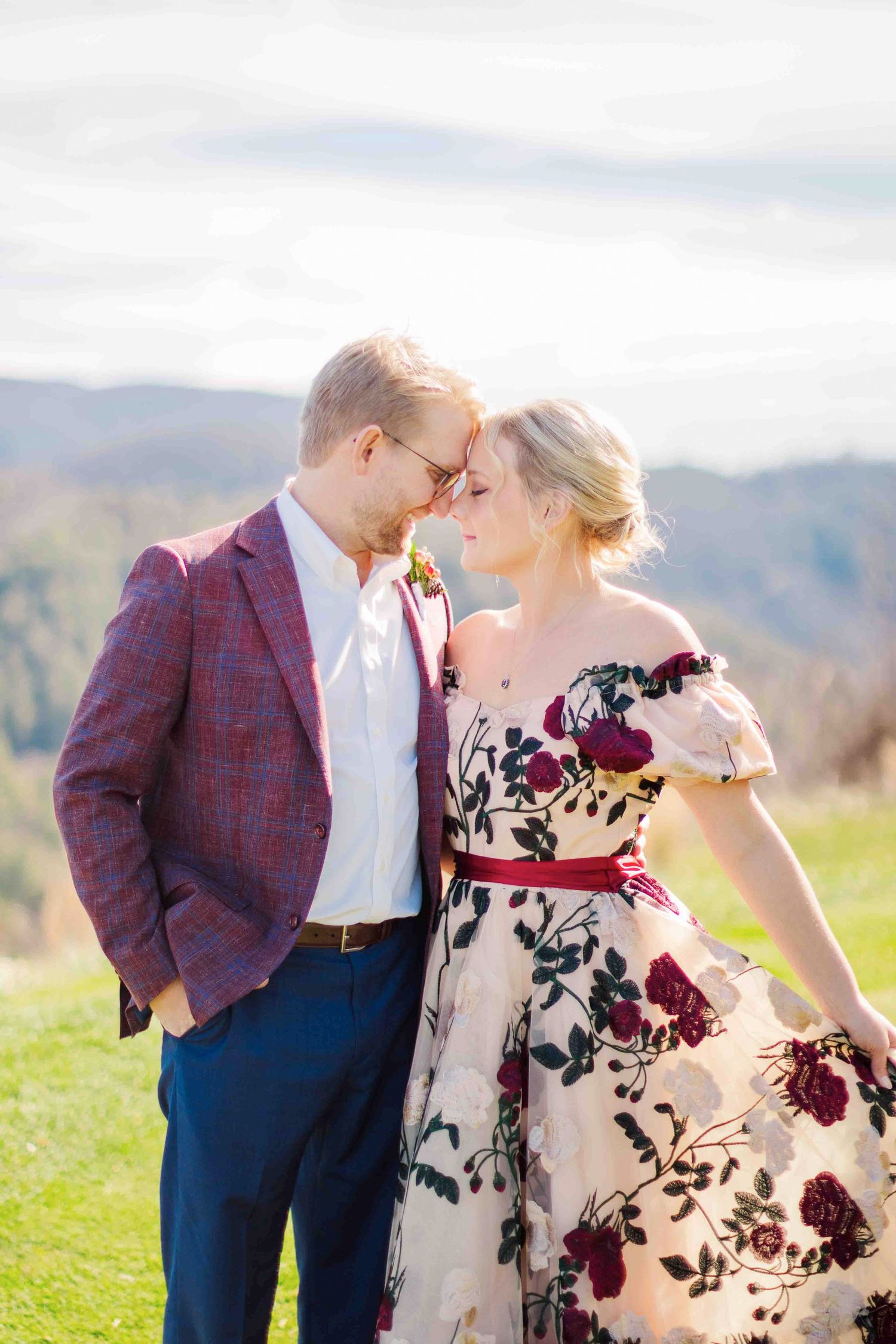 Couple shares a tender forehead touch in a floral wedding dress against a mountain landscape.