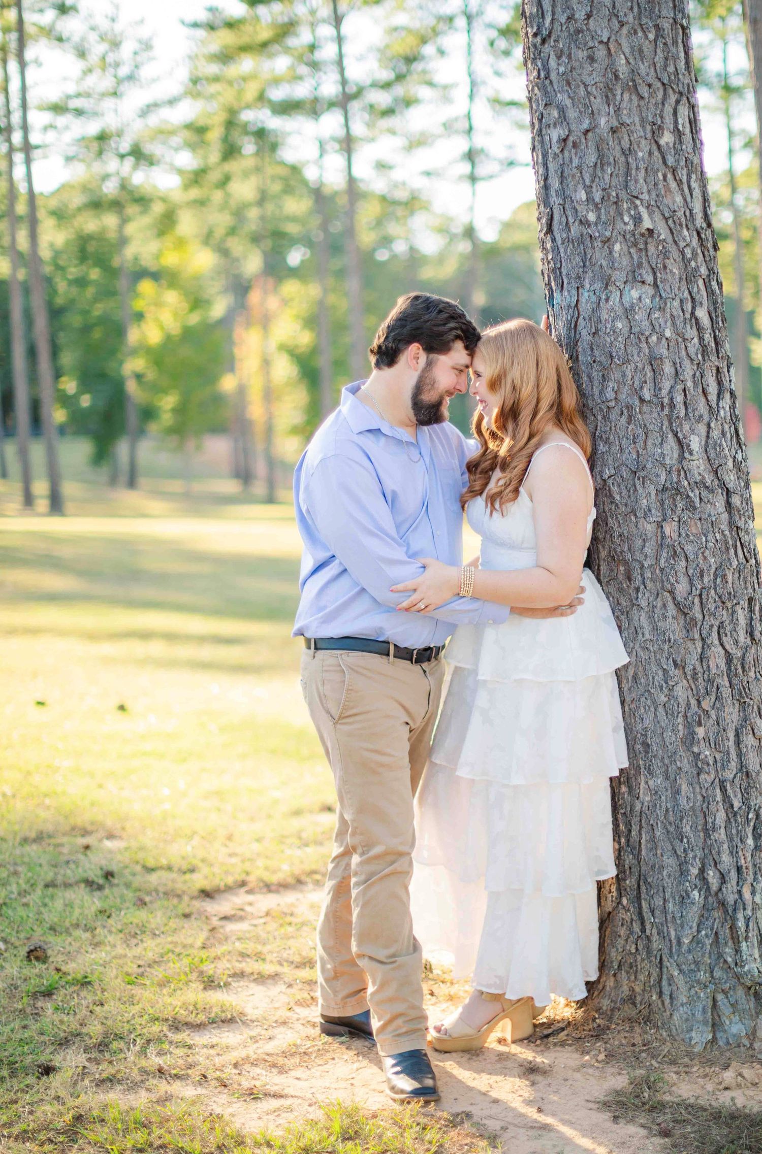 Couple leaning against pine tree holding hands, woman in white tiered dress, golden sunlight filtering through.