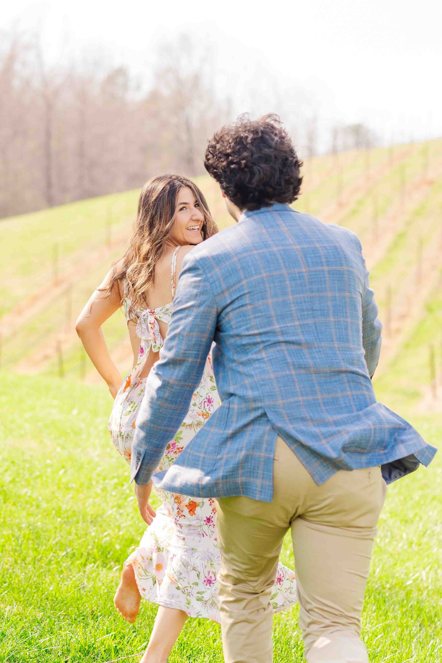 Couple dancing and laughing together in a sunny vineyard, woman in floral dress, man in blue plaid blazer.