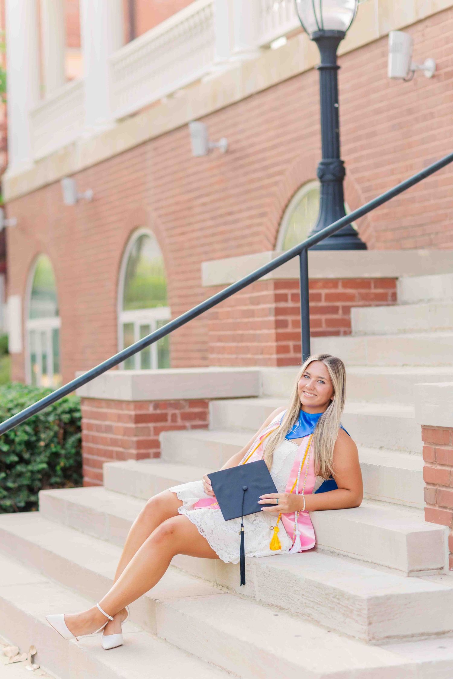 Blonde graduate in white dress sits on brick building steps holding cap with colorful stoles.
