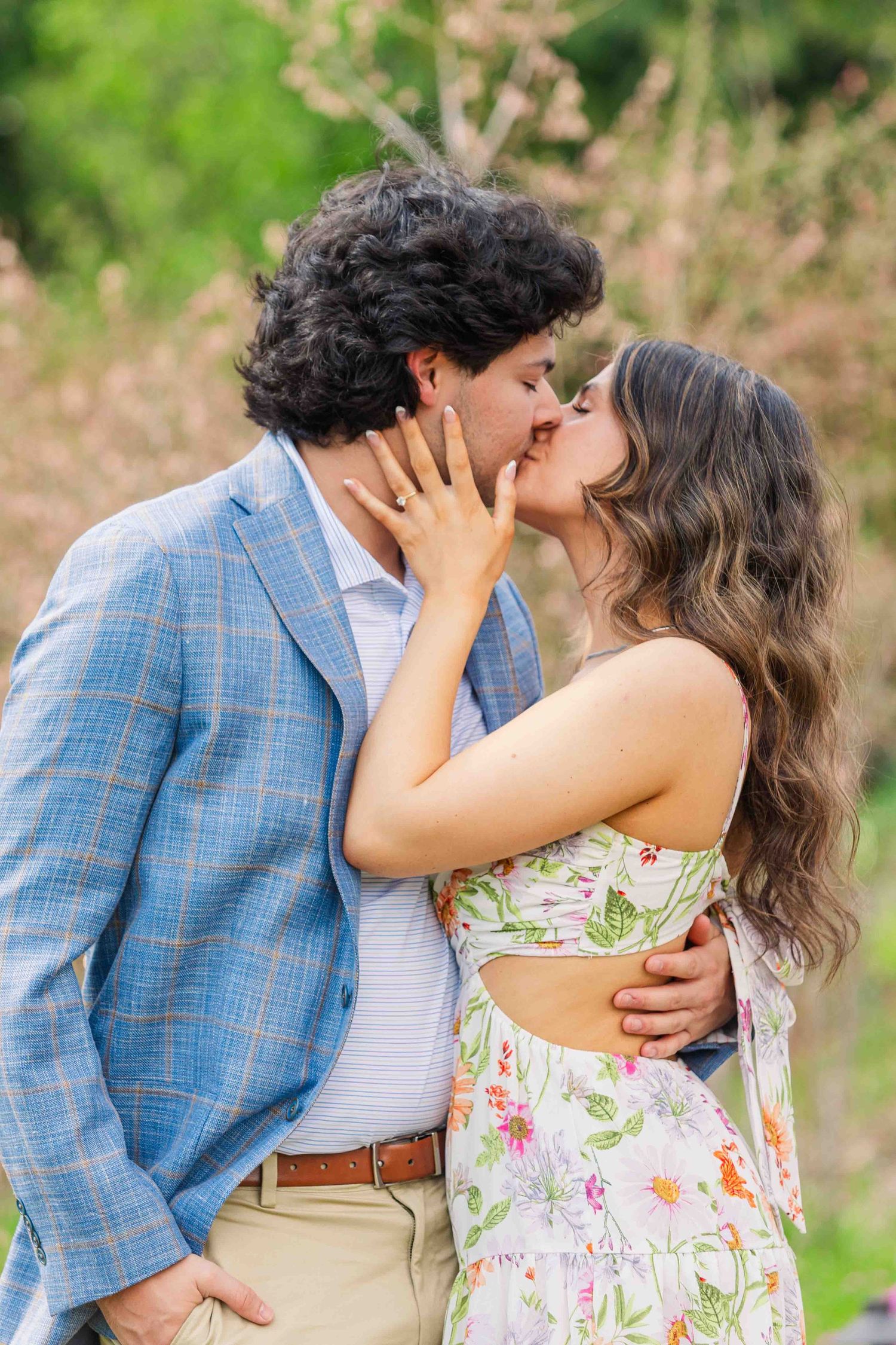 Couple sharing a romantic kiss outdoors, woman in floral dress touching man's face, green garden background.