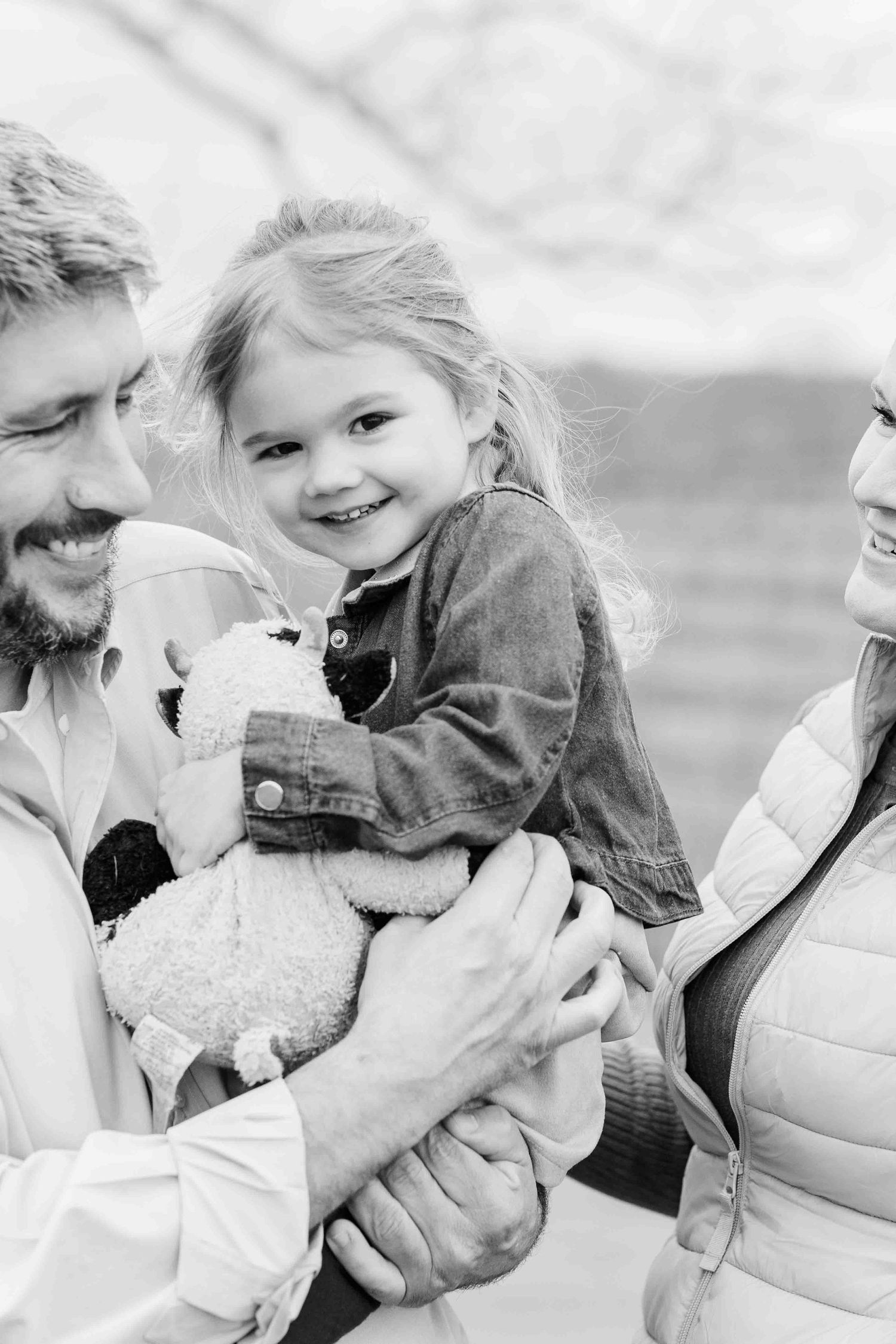 Black and white photo of smiling toddler girl held by parents outdoors, clutching a stuffed animal toy.