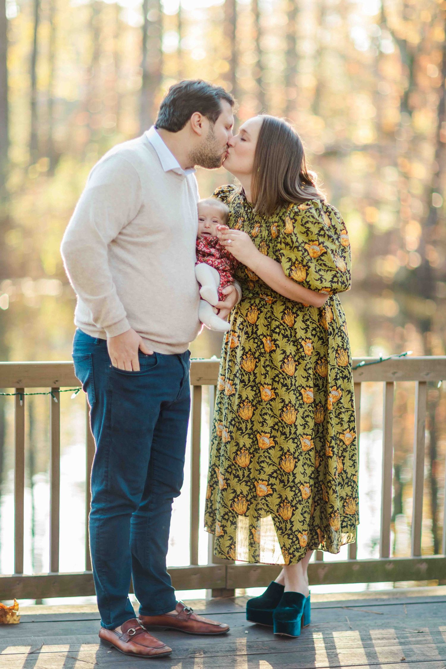 Couple kissing on wooden deck while holding newborn baby in autumn forest setting.
