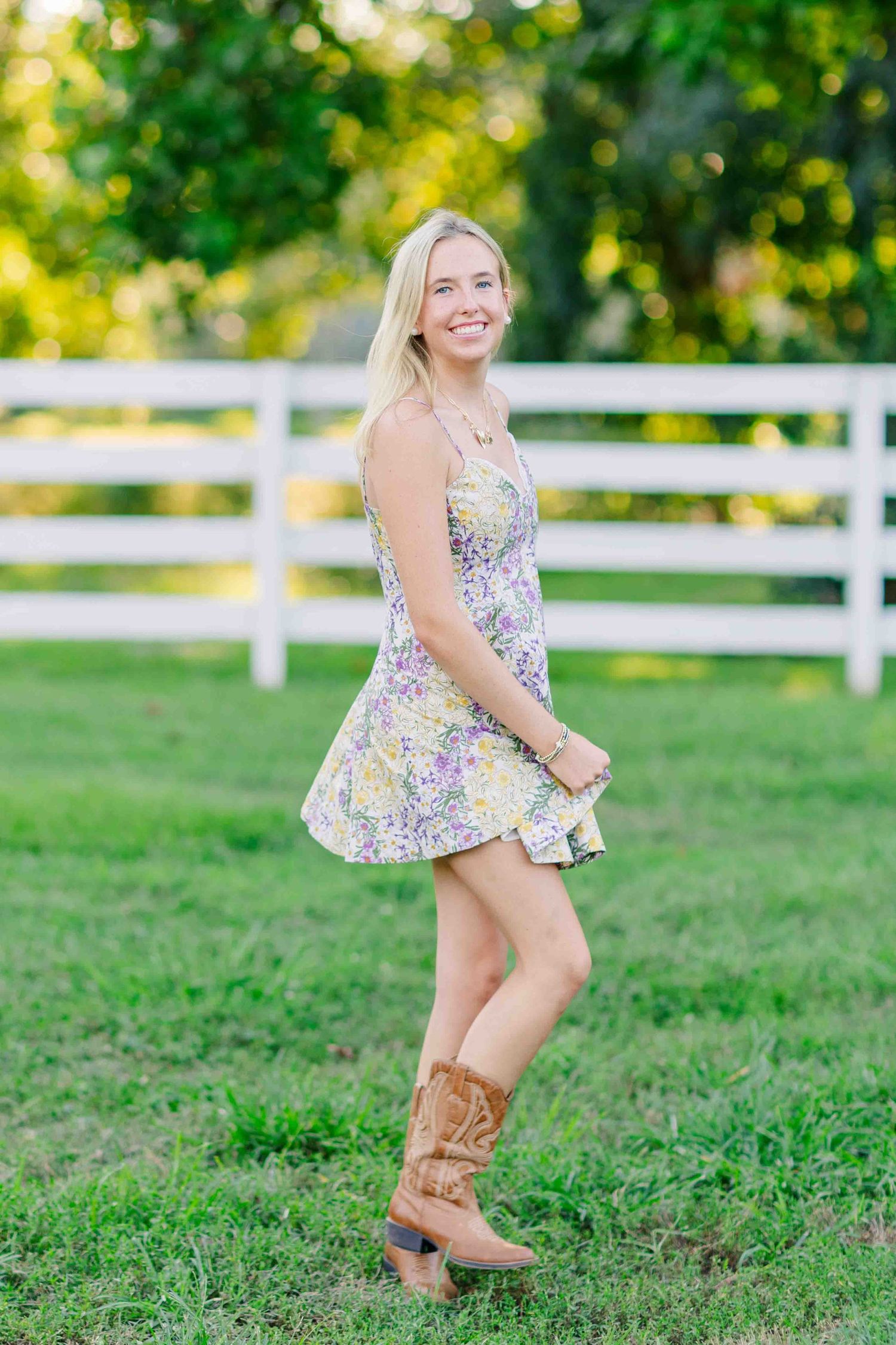 Smiling blonde woman in floral mini dress and cowboy boots posing near white fence outdoors.