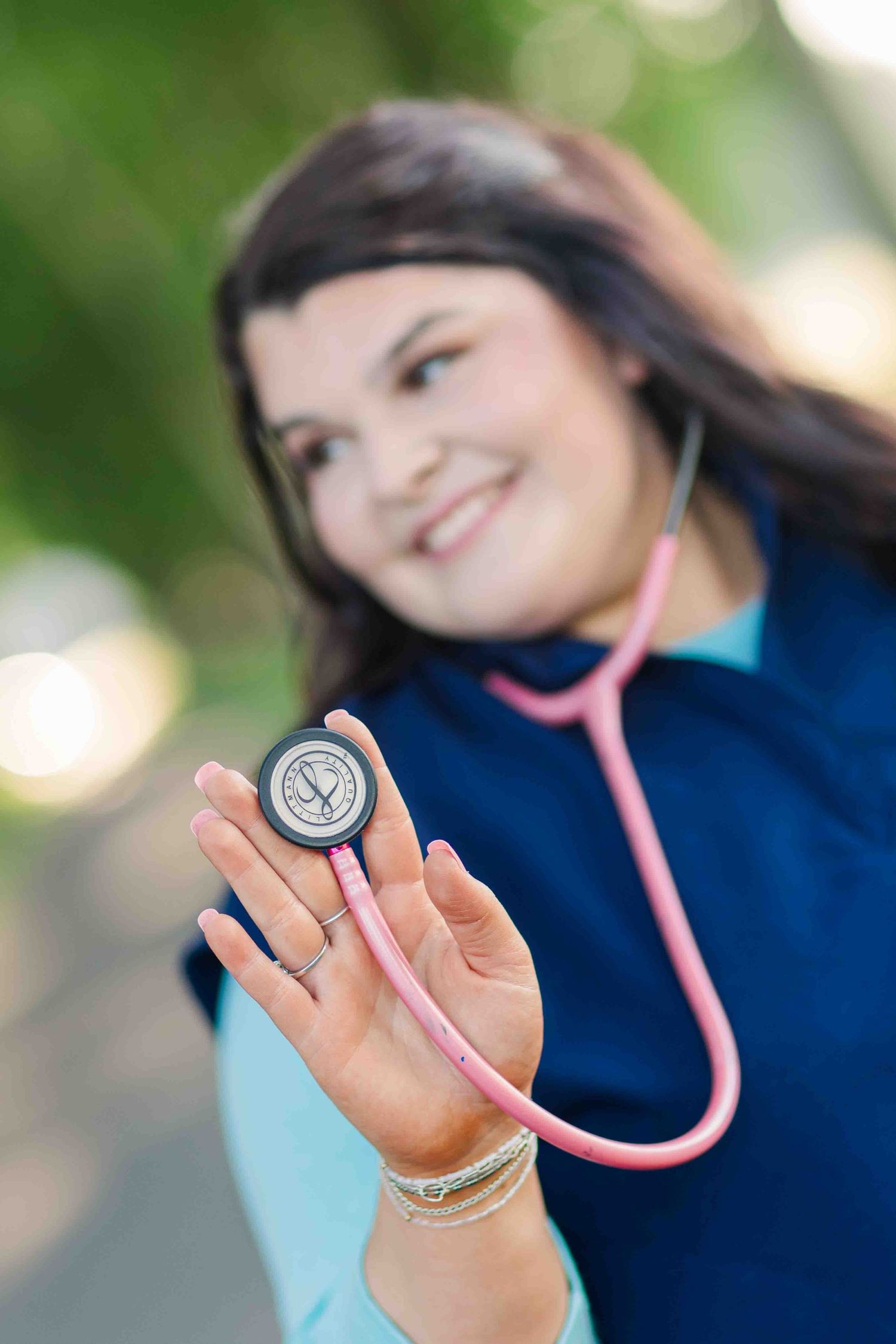 Smiling nursing student holding pink stethoscope toward camera in outdoor green setting.