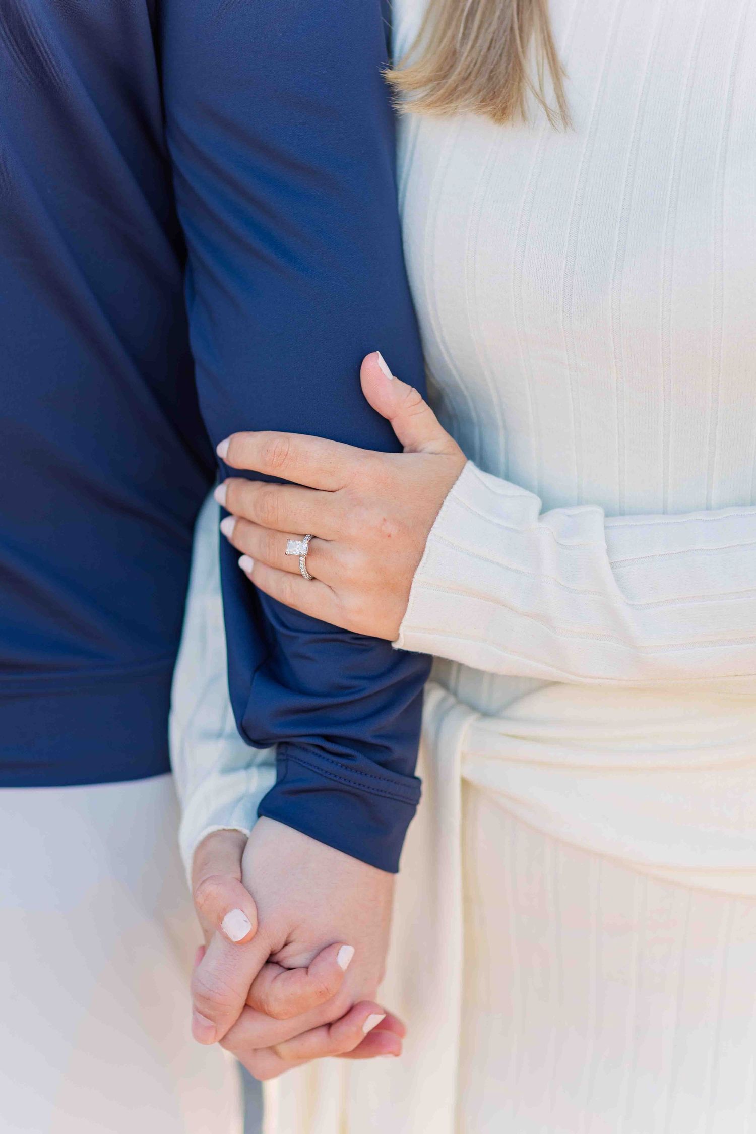 Close-up of womans hand with diamond engagement ring, holding partners hand while wearing cream ribbed dress.