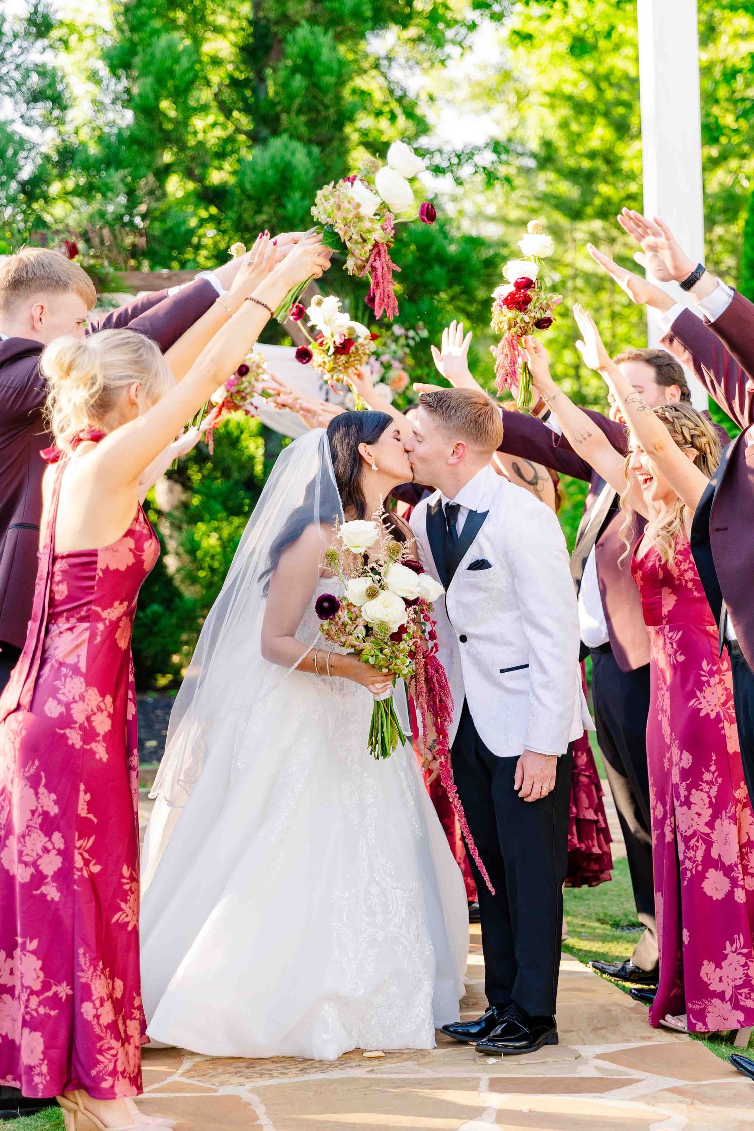 Bride and groom kissing under raised bouquets as wedding party in burgundy dresses and white suits celebrate outdoors.