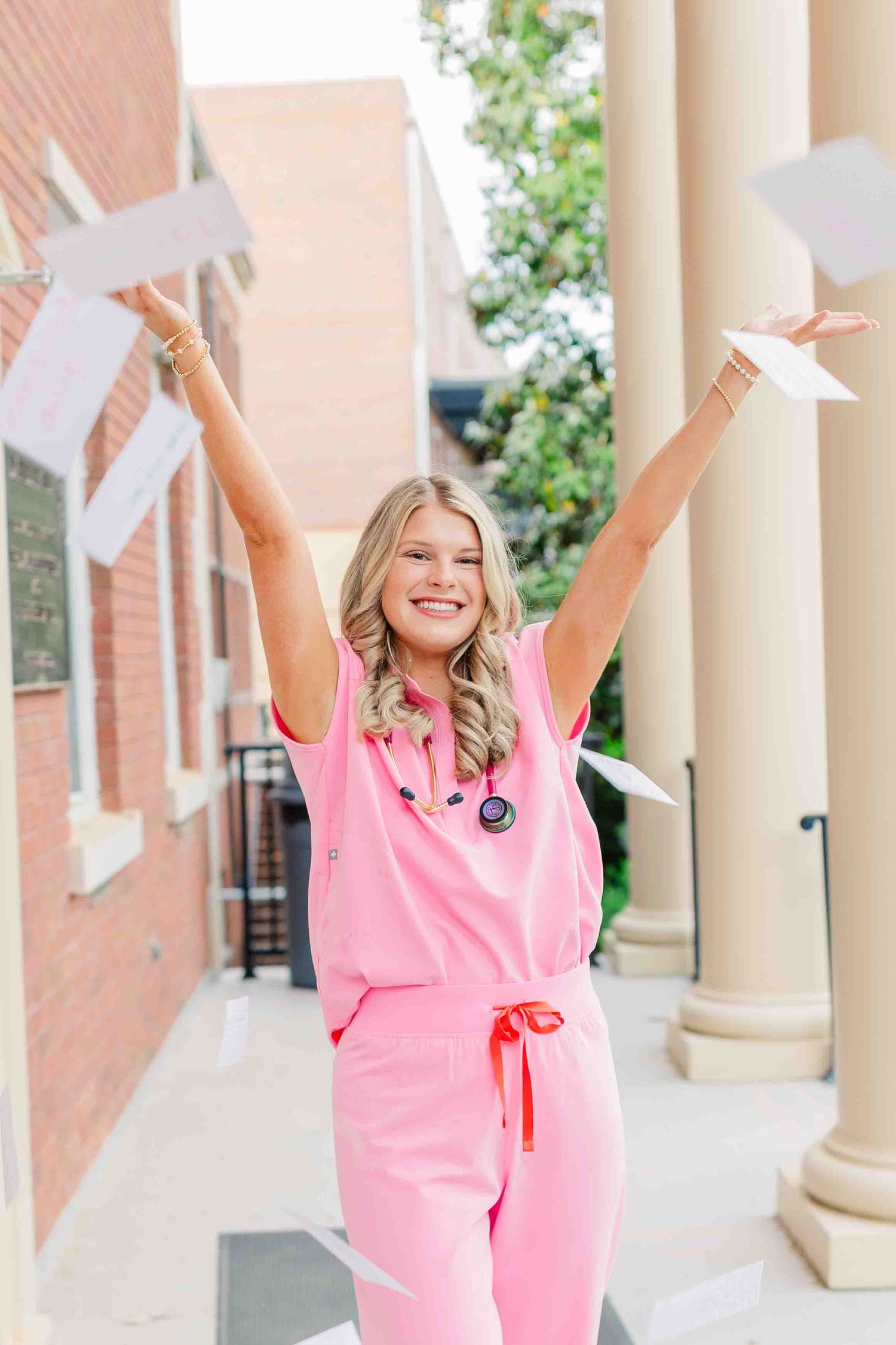Smiling blonde woman in pink scrubs with stethoscope tossing papers in the air outdoors.