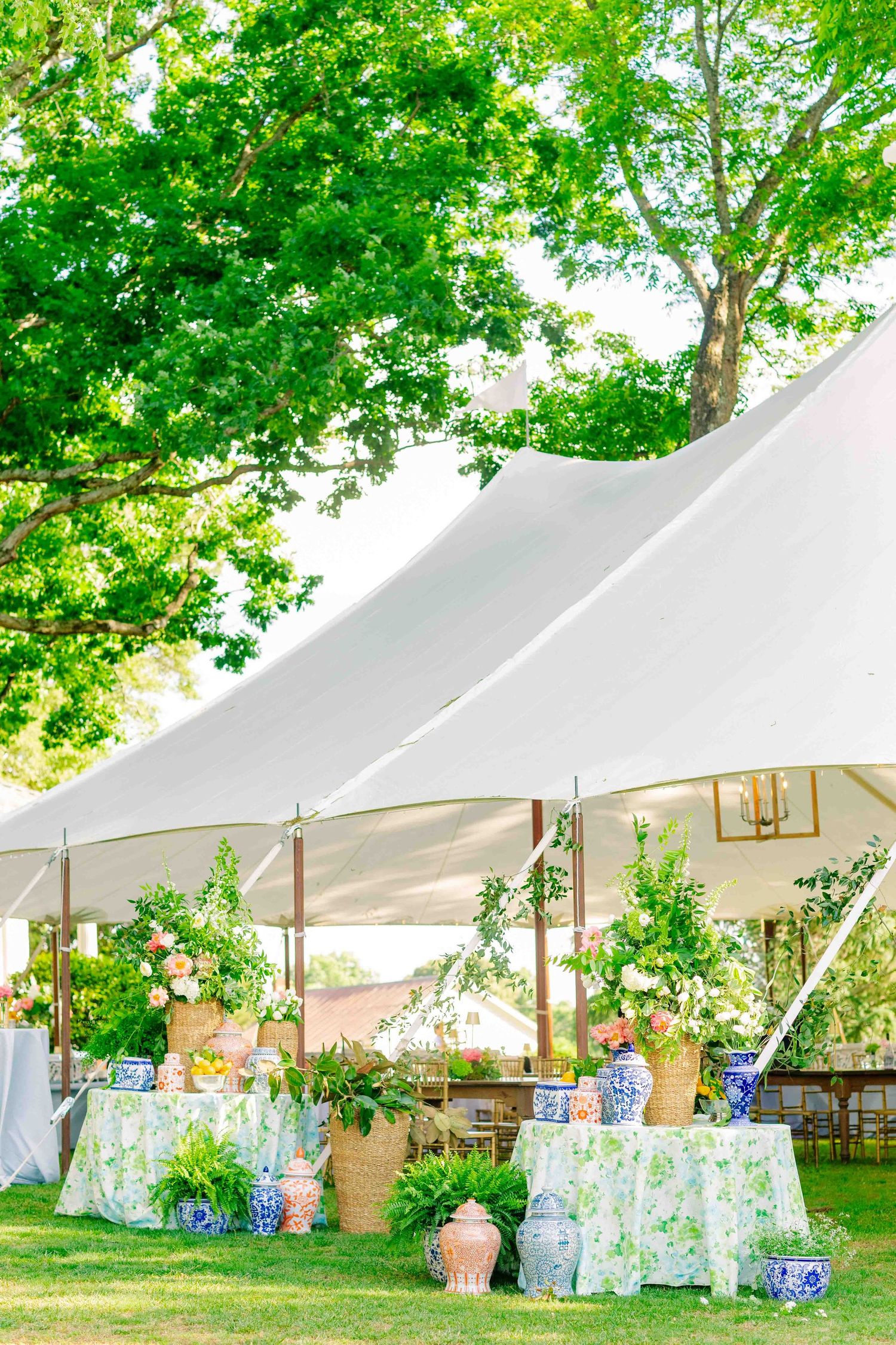 Elegant outdoor wedding reception tent with blue and white floral arrangements, green topiaries, and floral tablecloths on a sunny day.