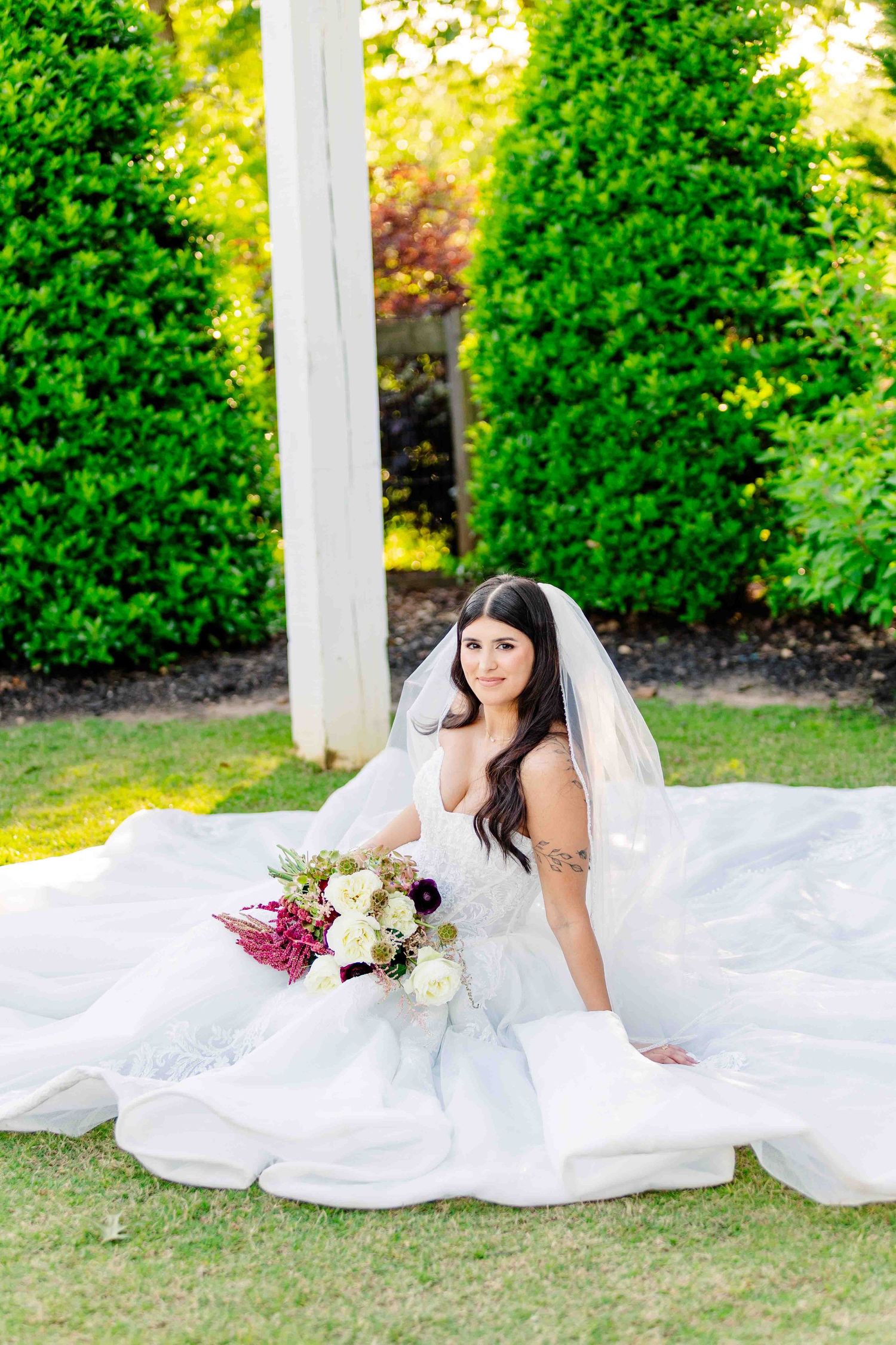 Brunette bride in flowing white gown and veil seated on lawn with floral bouquet.