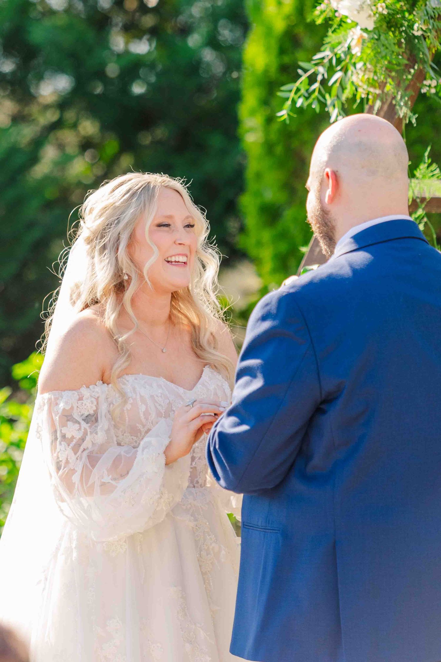 Blonde bride in lace off-shoulder gown smiles at groom during outdoor wedding ceremony.