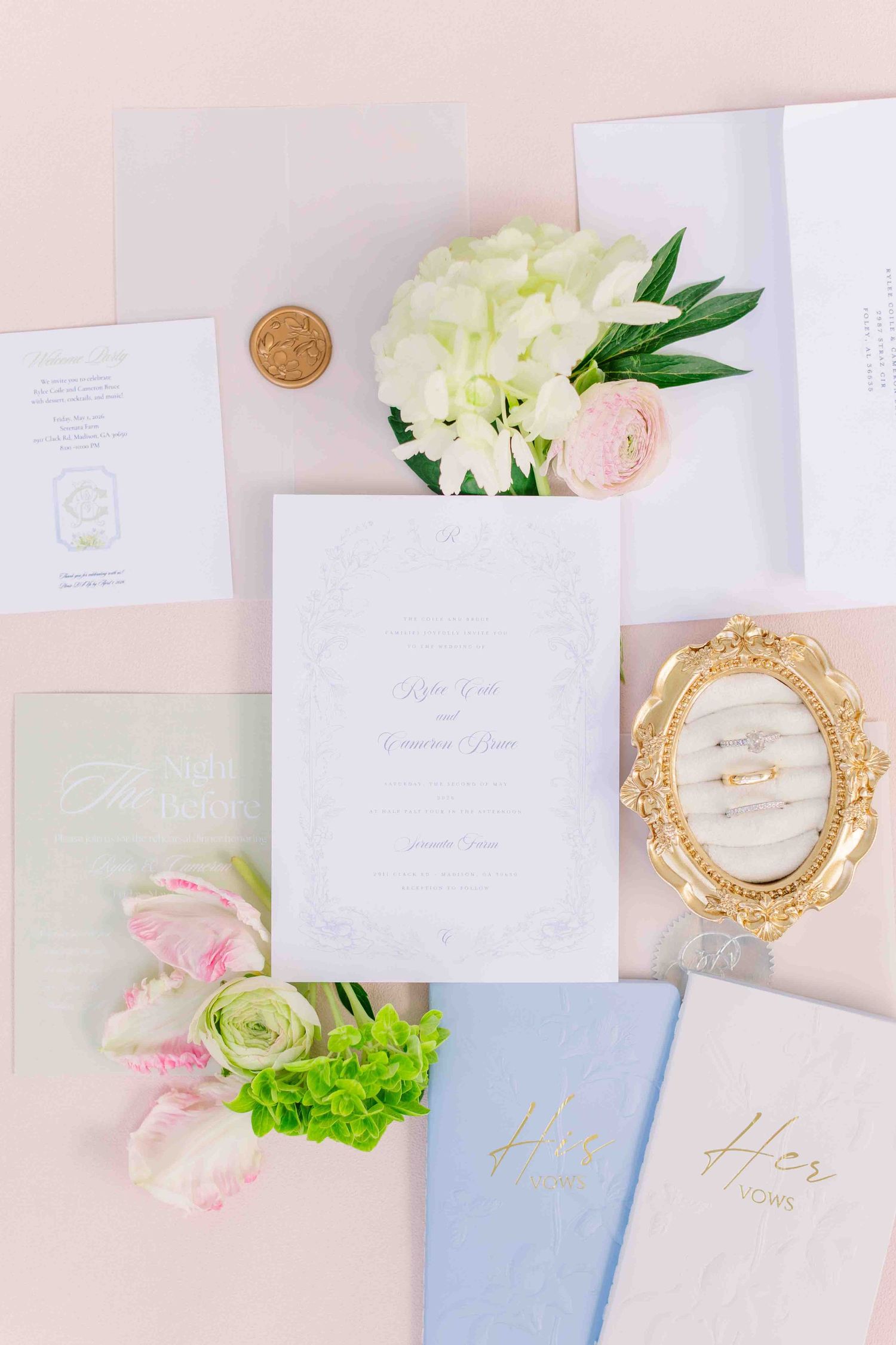 Wedding stationery flatlay with invitations, rings, flowers, and gold wax seal on pink background.