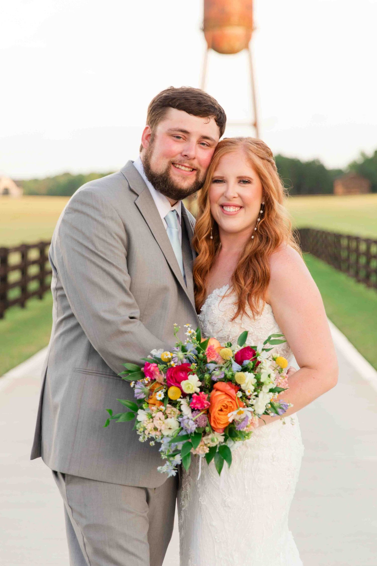Smiling bride and groom pose with a colorful bouquet near a rustic water tower at sunset.