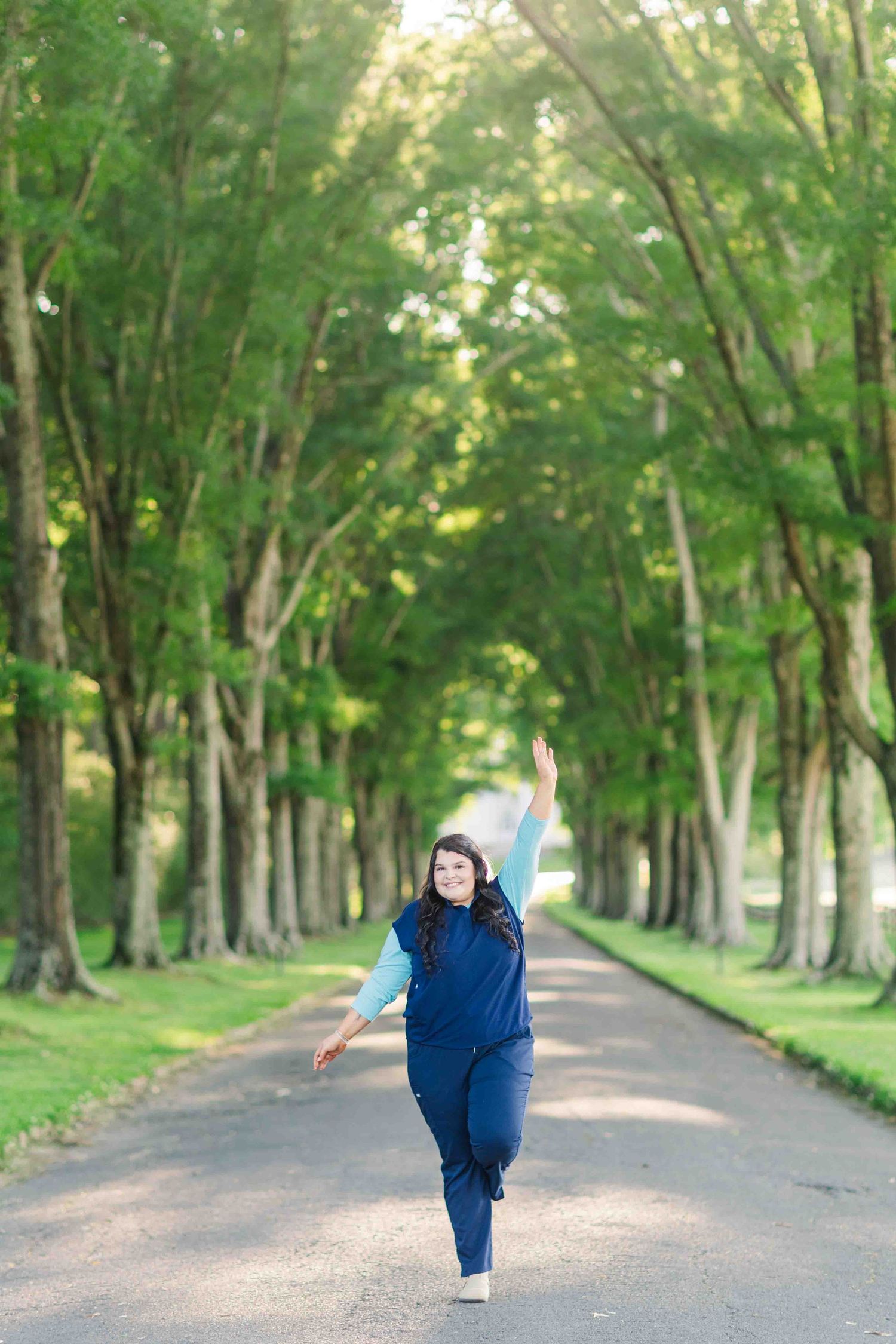 Nursing student in blue scrubs joyfully walking down tree-lined path on sunny day.
