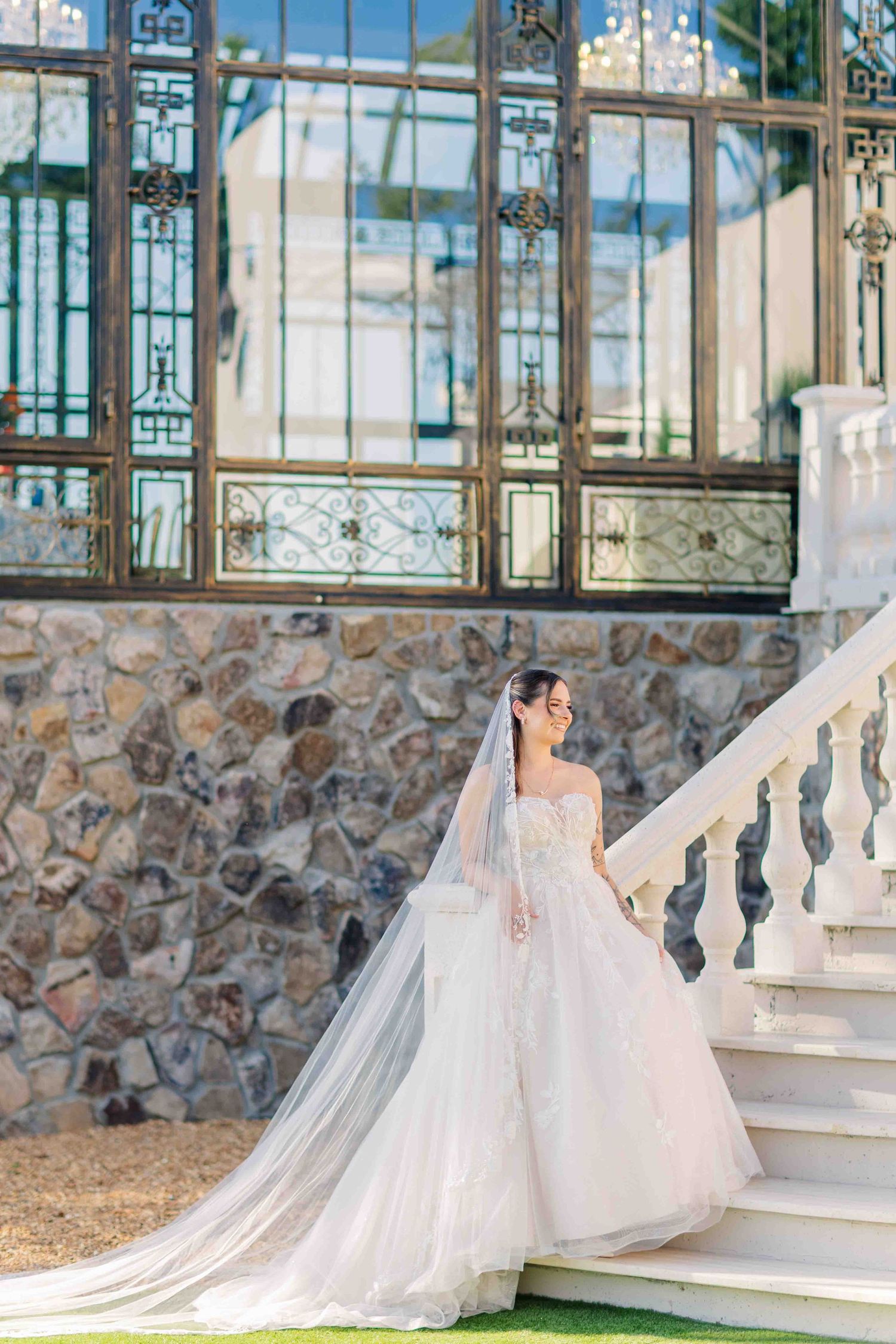 Bride in a flowing white lace gown and veil stands on elegant white stairs outside a stone building.
