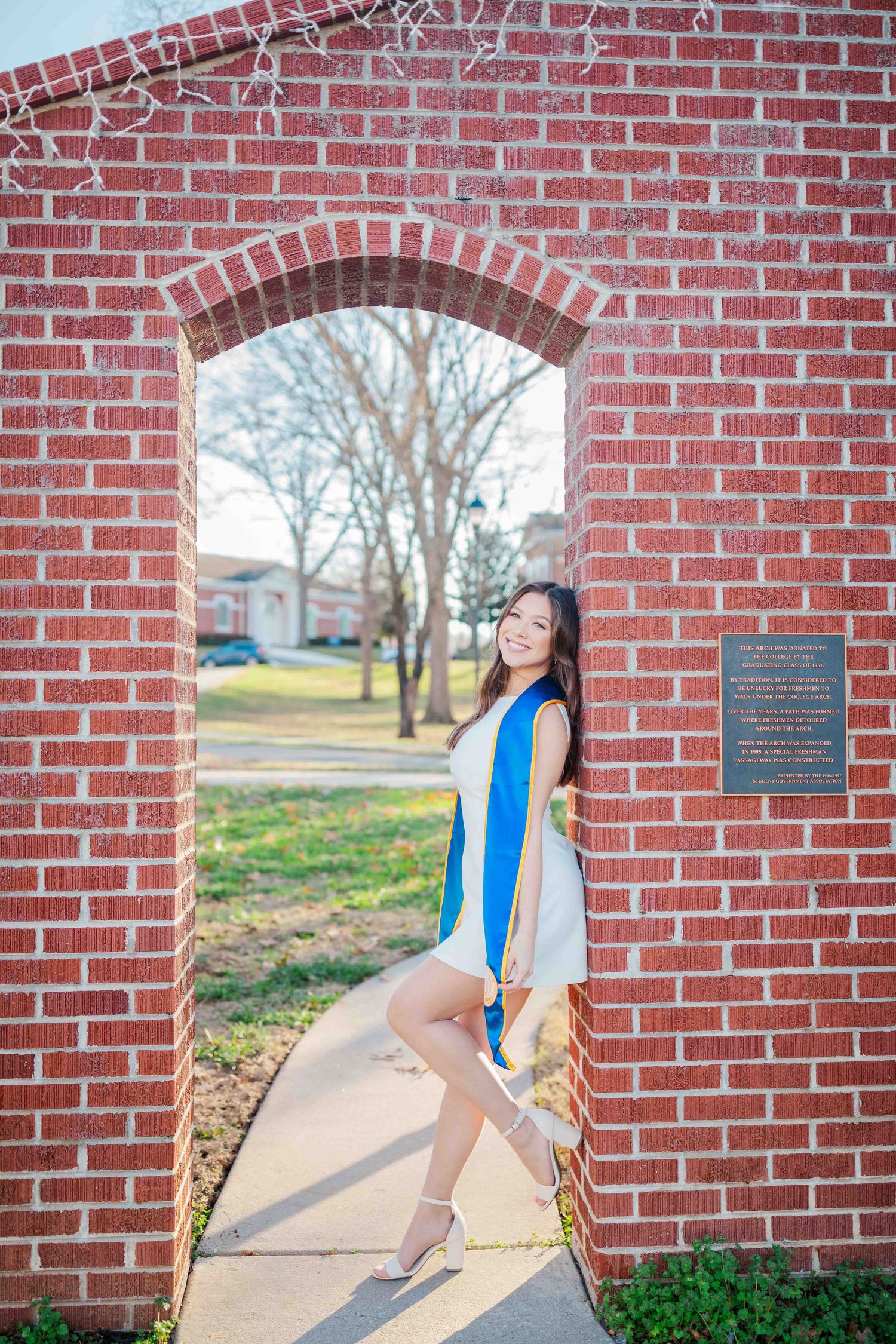 Young woman in white dress and blue graduation sash poses in brick archway on college campus.