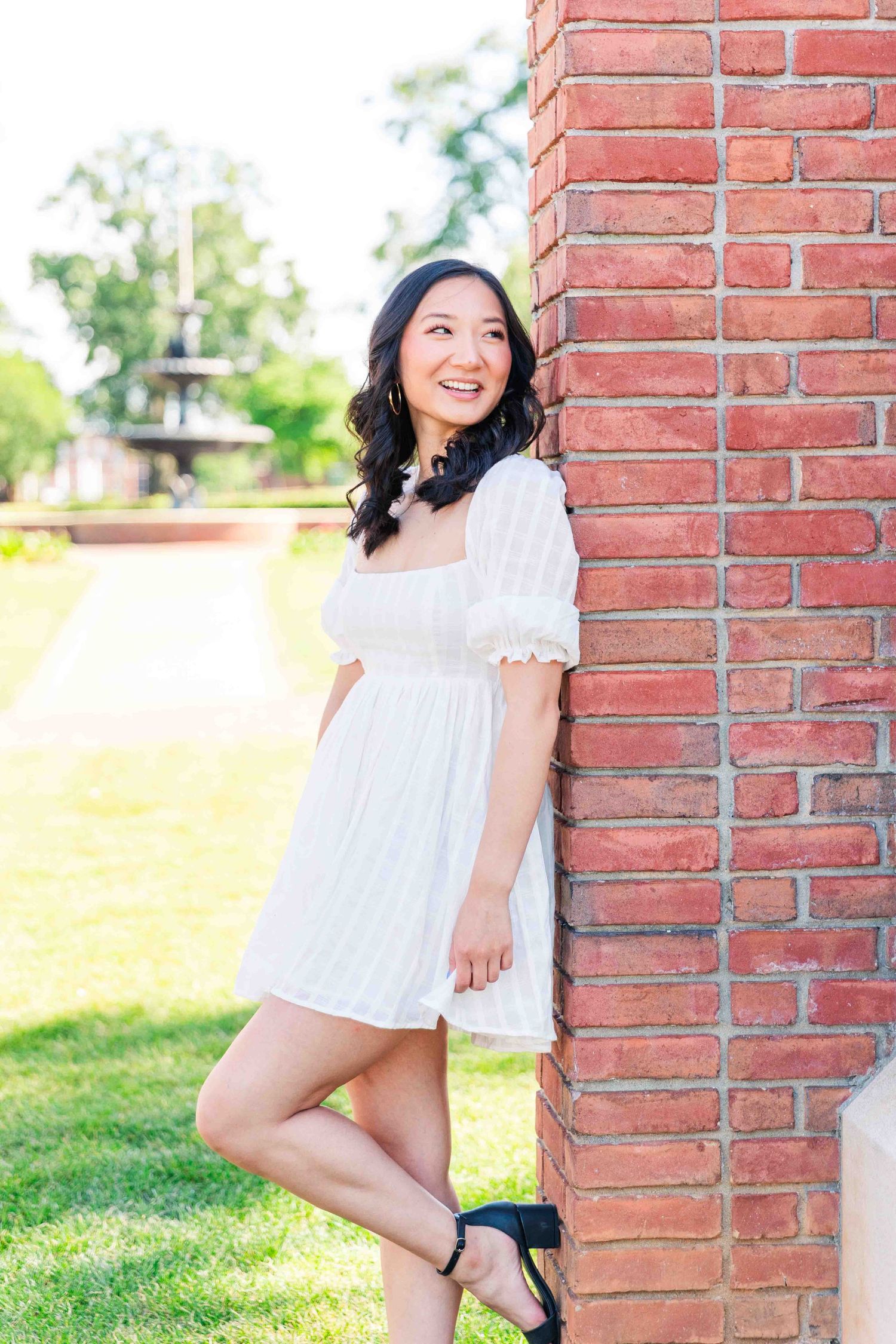 Smiling young woman in white dress leans against brick wall on sunny campus grounds.
